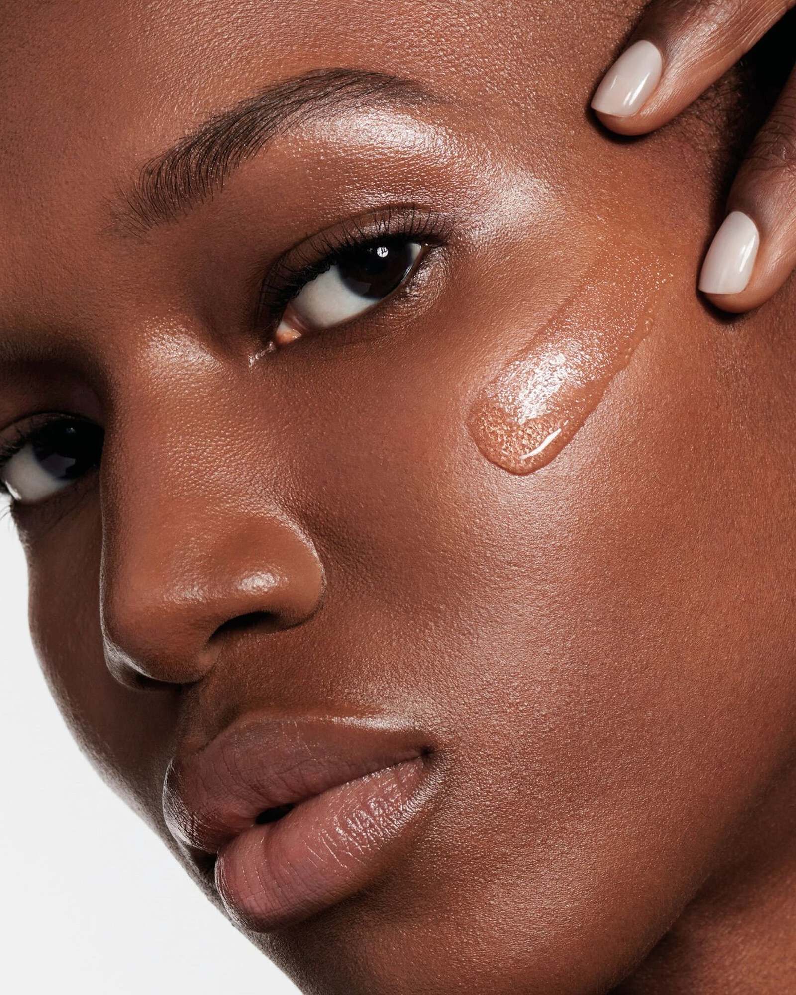 Extreme close-up of a woman with deep brown skin and a glossy swipe of liquid foundation on her cheek