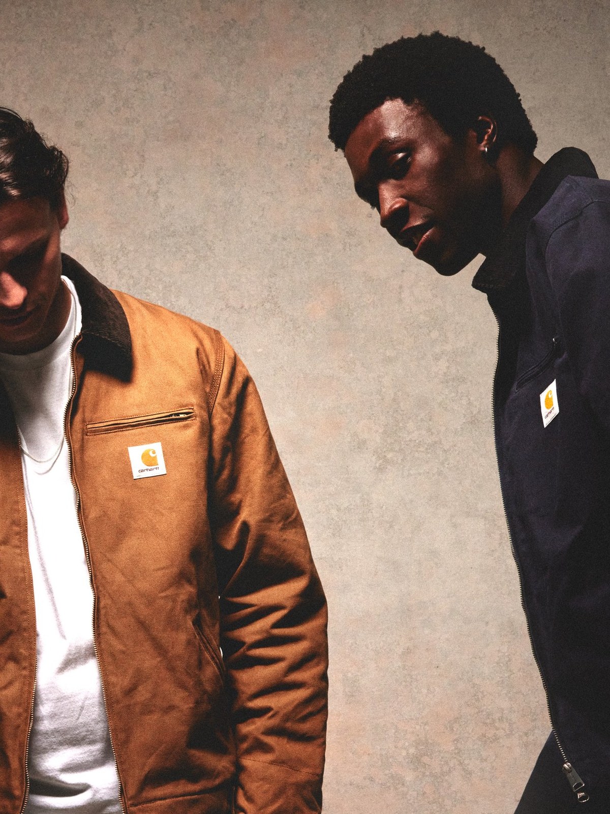 Two male models in tan and navy Detroit jackets against a plaster wall