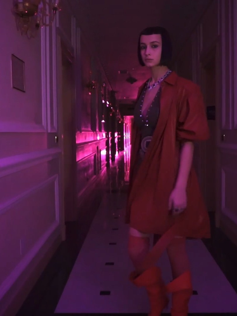 Woman in a rust-red coat and dress in a pink-lit hotel hallway