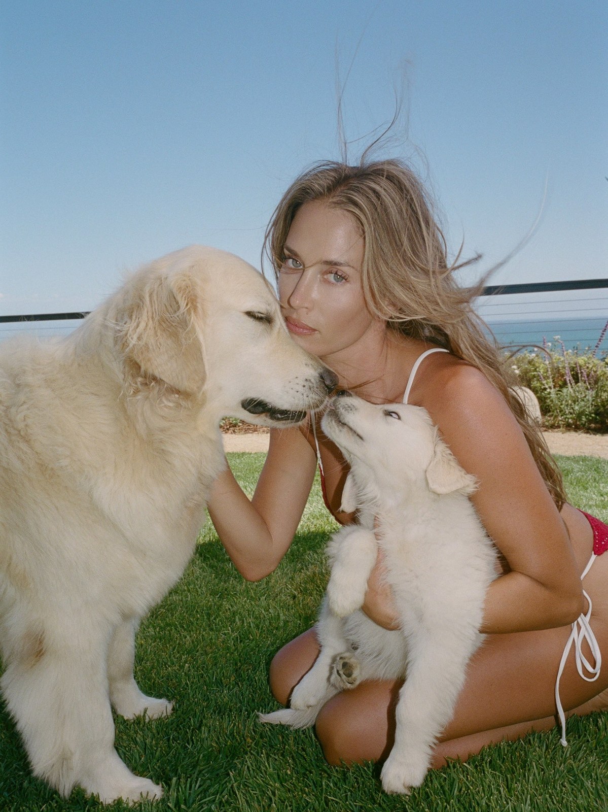 Woman in a red bikini outdoors beside two dogs near the ocean