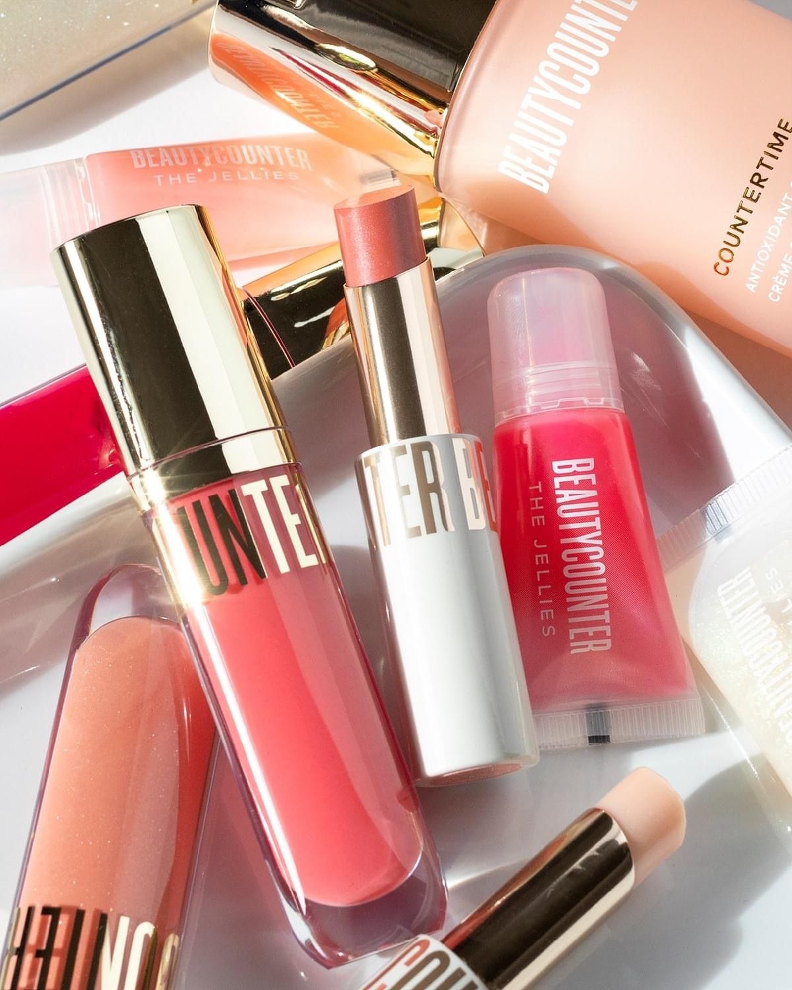 Flat lay of pink lip glosses, a lipstick, a jelly tube and a peach moisturizer bottle with gold caps on a glossy white surface