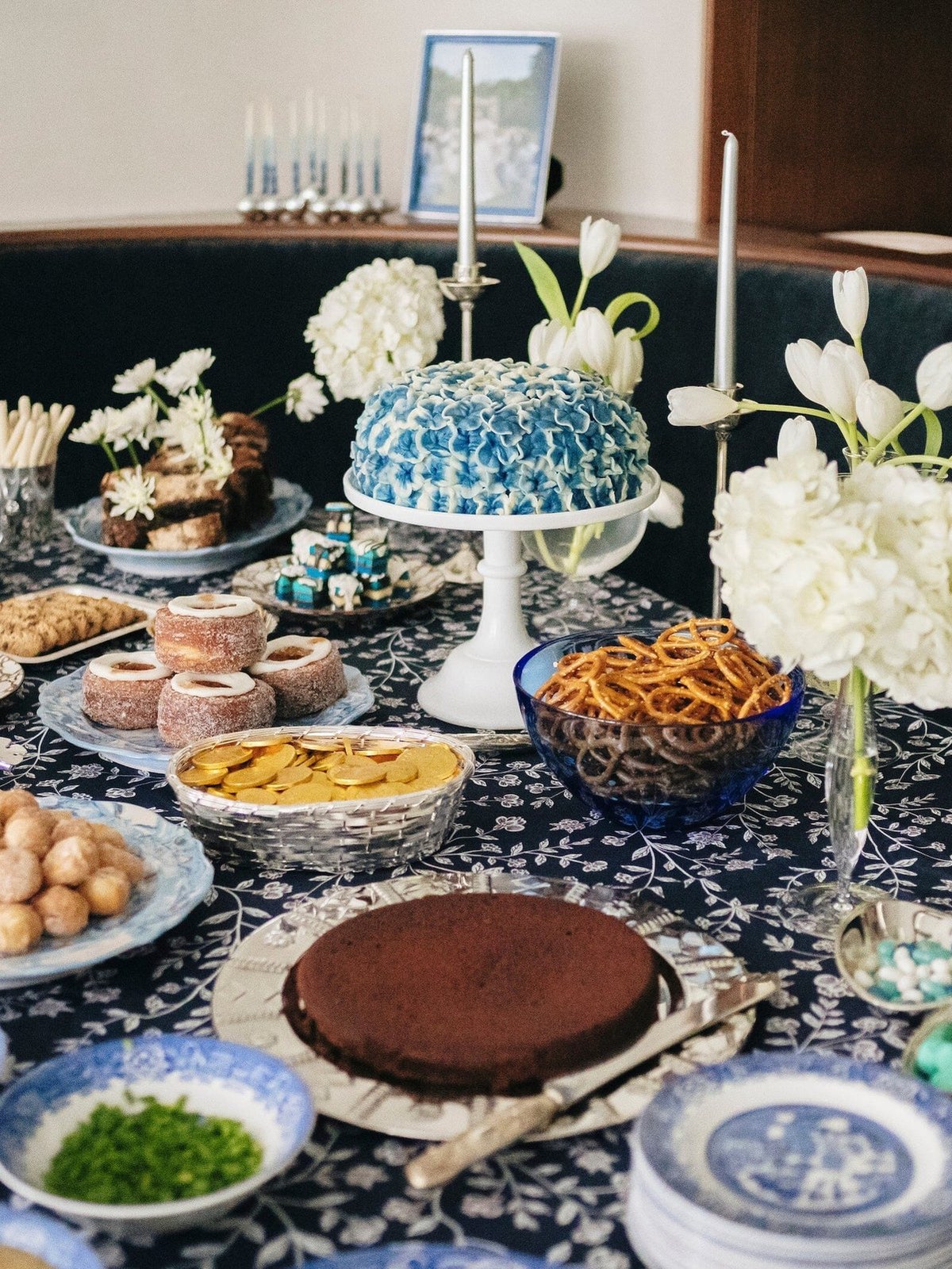 Chanukah dessert table dressed in dark patterned linens with flowers, cakes, and blue and white plates