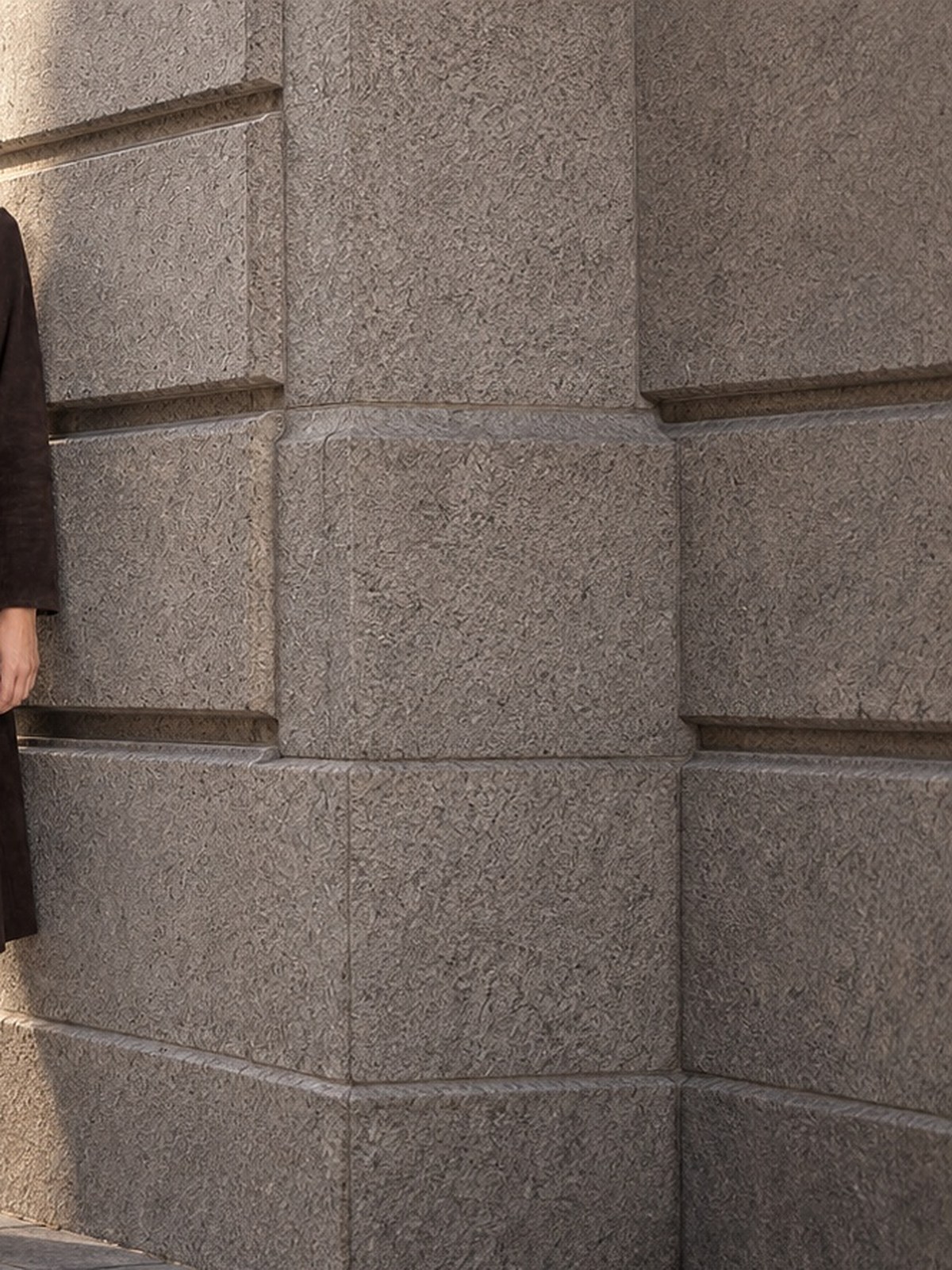 Model in a long dark suede coat and white trousers by a granite building