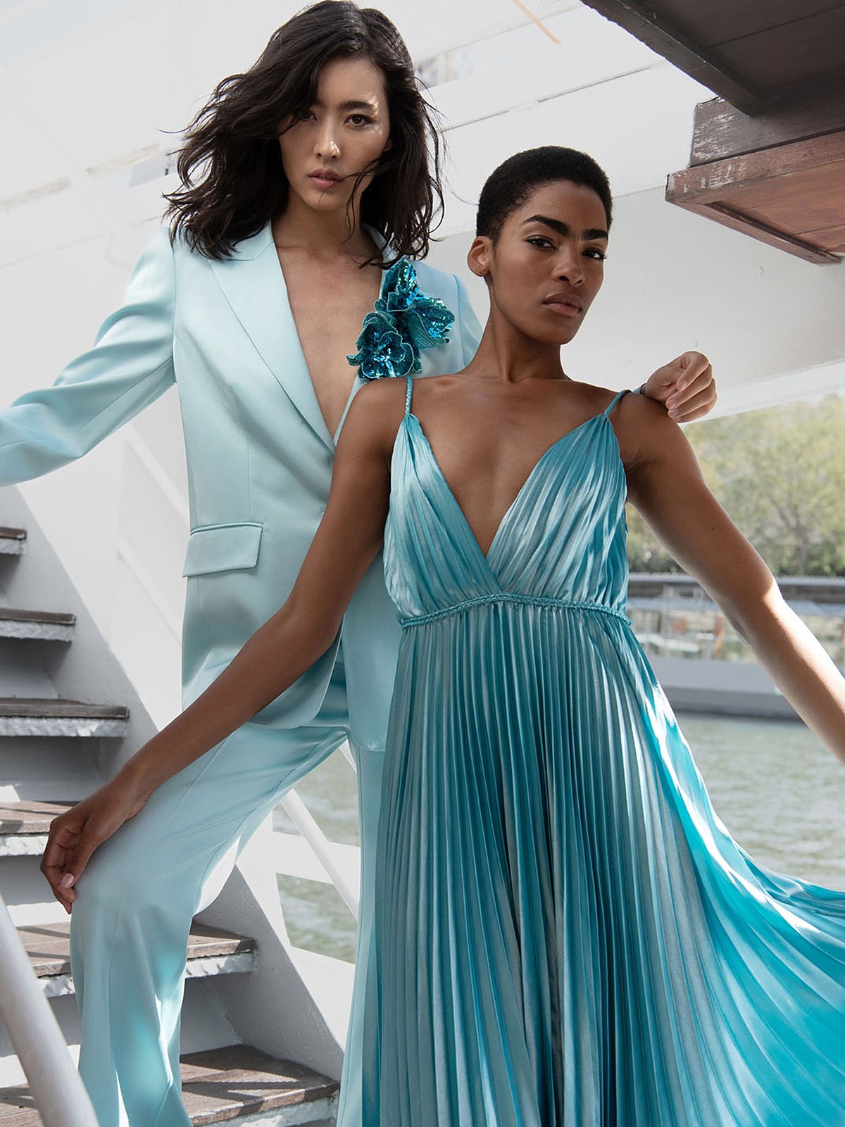 Two models in an aqua satin pantsuit and pleated turquoise gown on a boat staircase