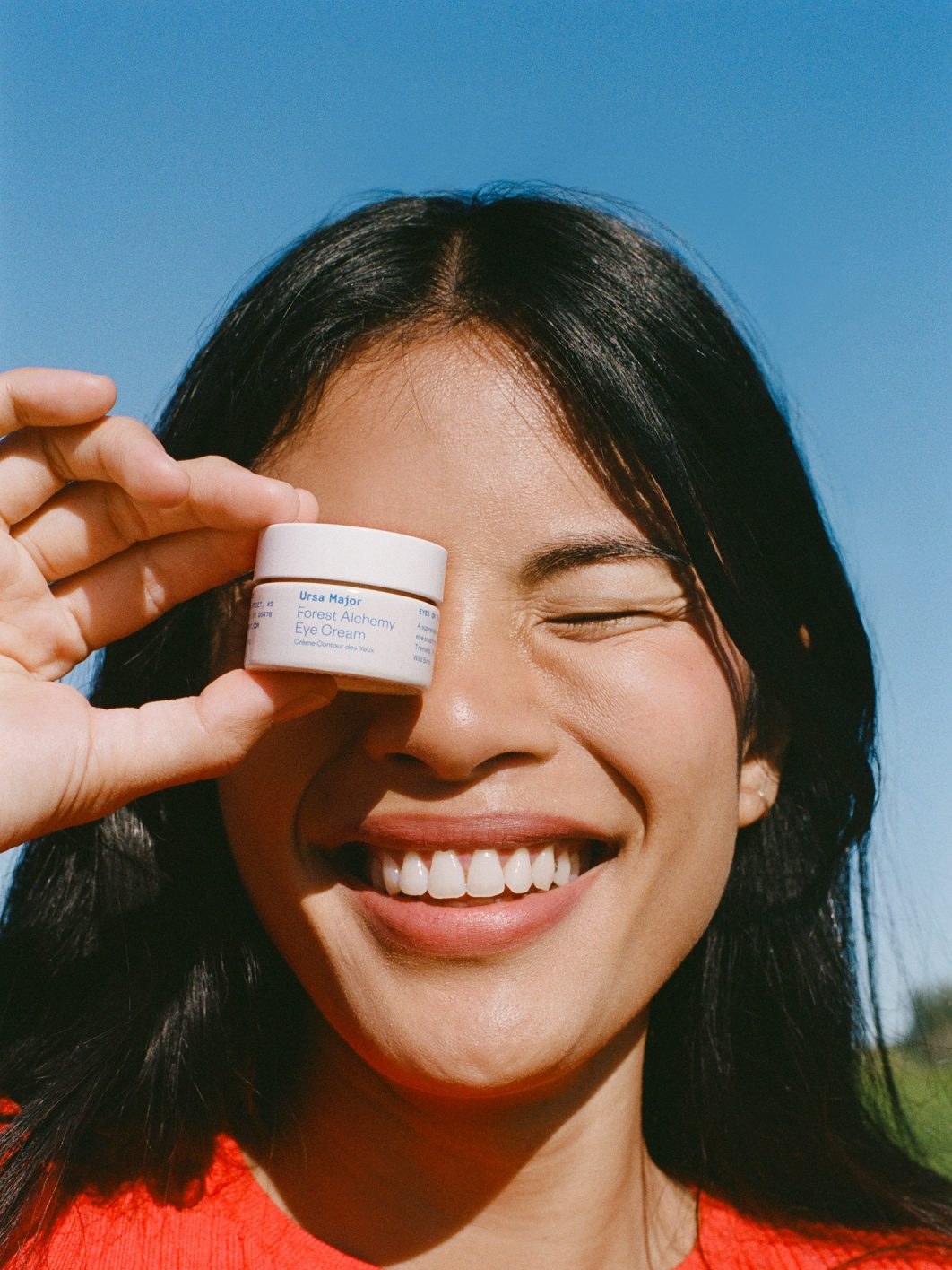 Woman laughing while holding a jar of Ursa Major eye cream in bright sunlight
