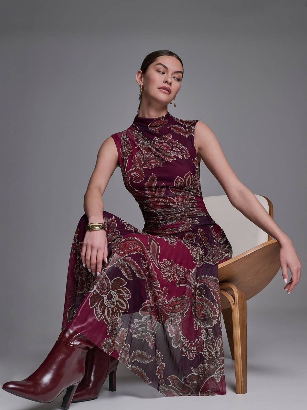 Model seated on a chair in a burgundy paisley mesh dress and oxblood boots