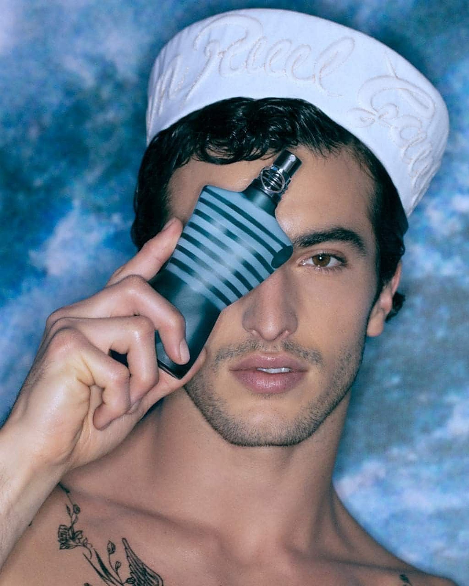 Dark-haired man in a white embroidered sailor cap holding a striped Jean Paul Gaultier torso bottle over one eye against a blue cloudy backdrop