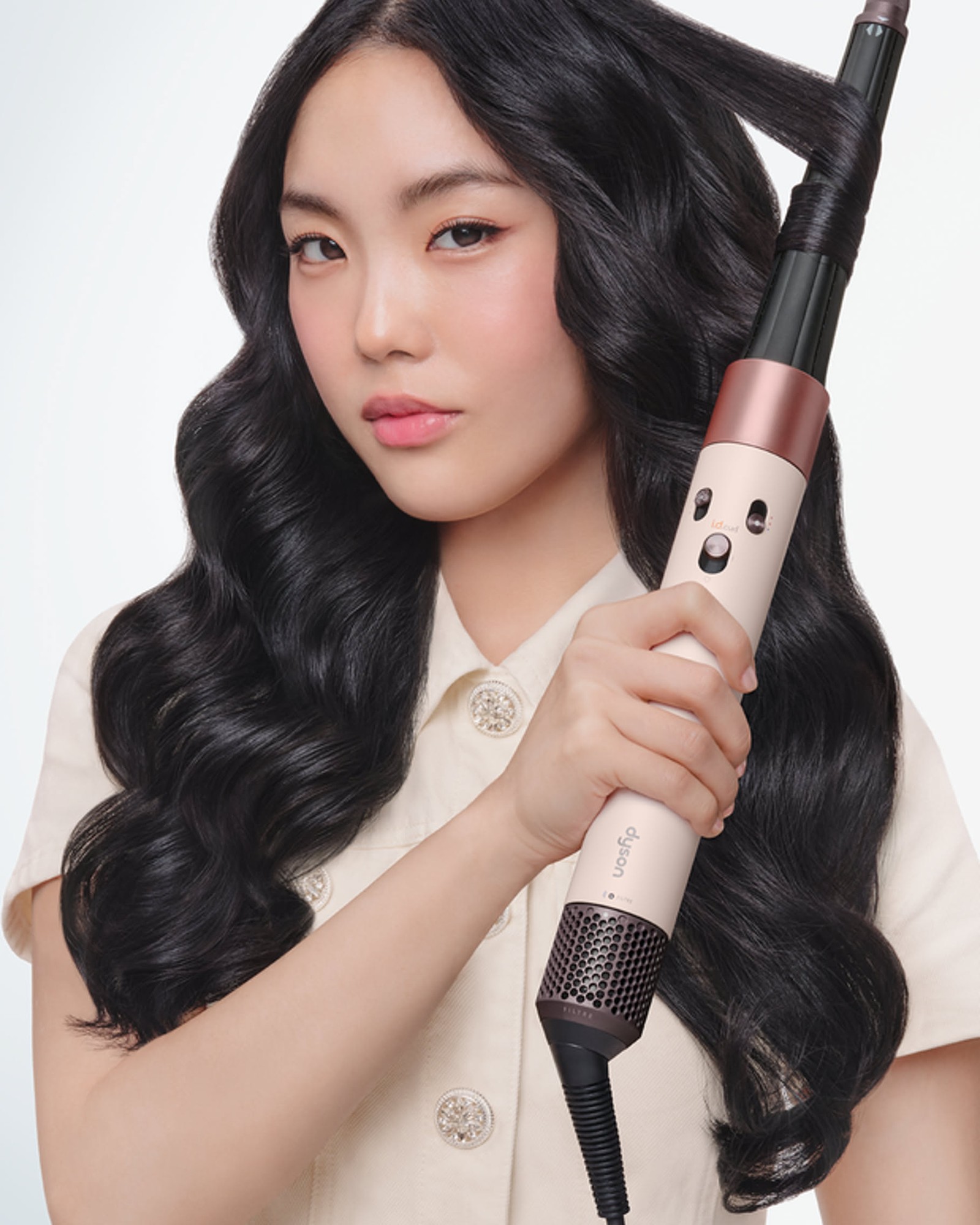 Woman with long dark wavy hair wrapping a lock around a pale pink and rose gold Dyson styling tool, wearing a cream button-up top