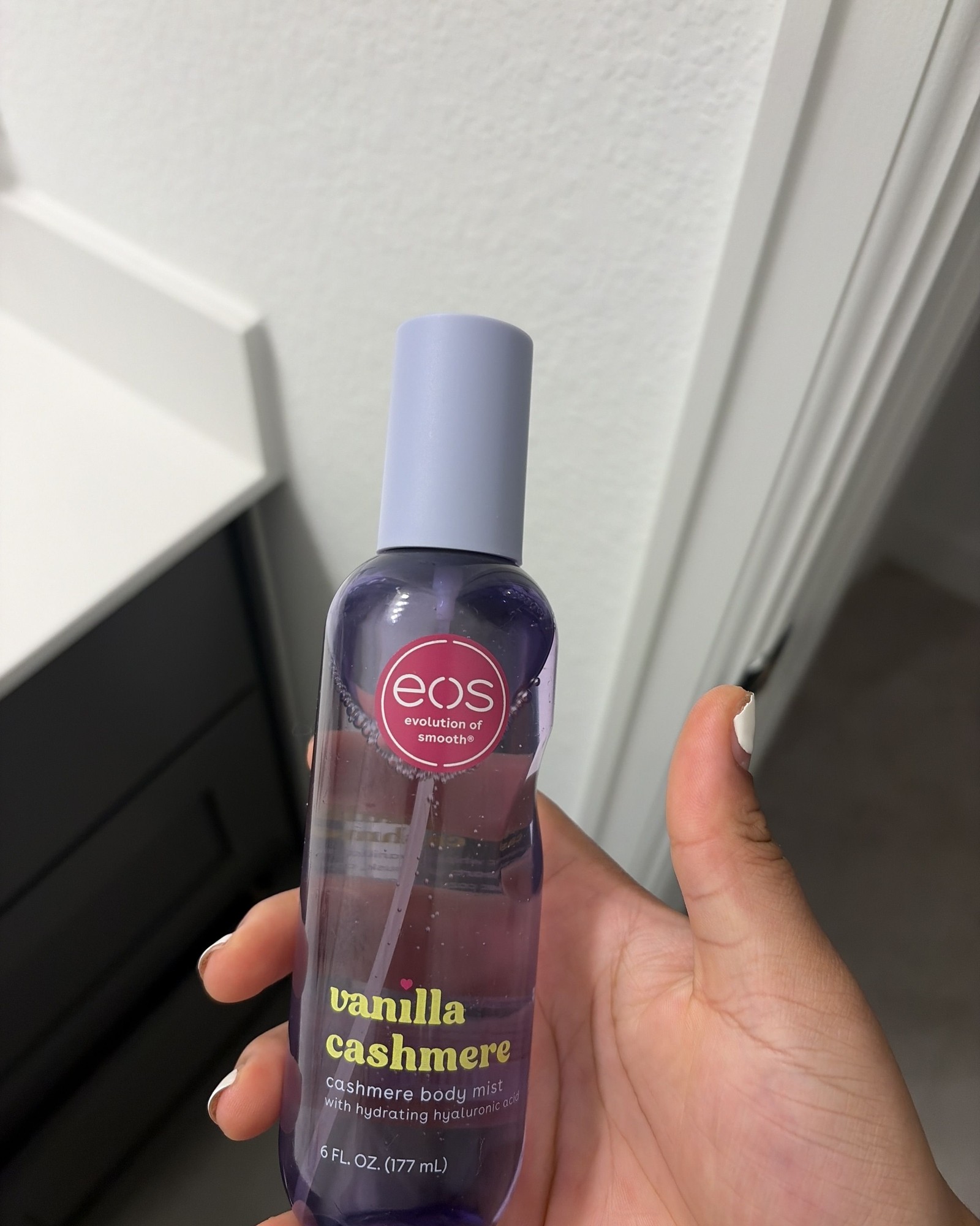 Hand holding an eos Vanilla Cashmere body mist bottle with a lavender cap in front of a white wall