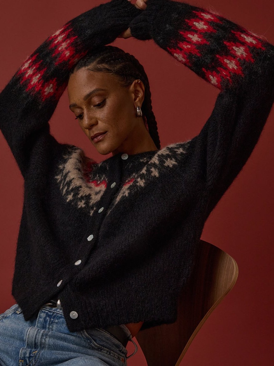 Model in a patterned cardigan and denim raising her arms against a red studio backdrop