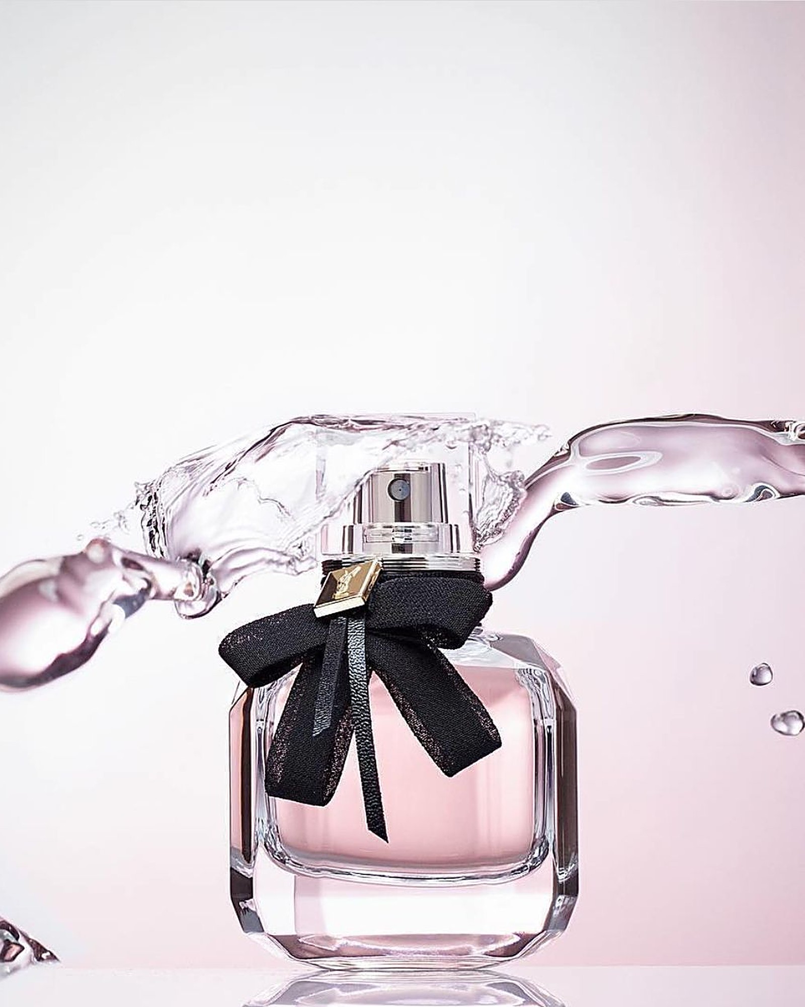 YSL Mon Paris bottle with its black satin bow and gold YSL tag, with a ribbon of water splashing across it against a soft pink background.