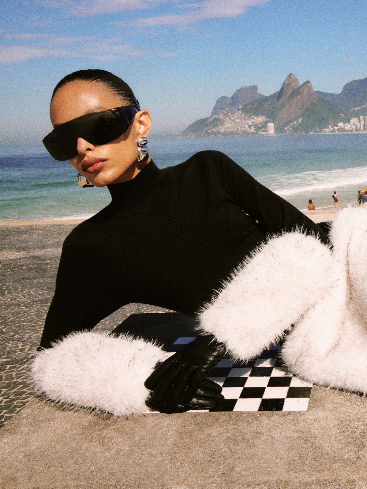Model reclining on a beach in a black top with white faux-fur sleeves
