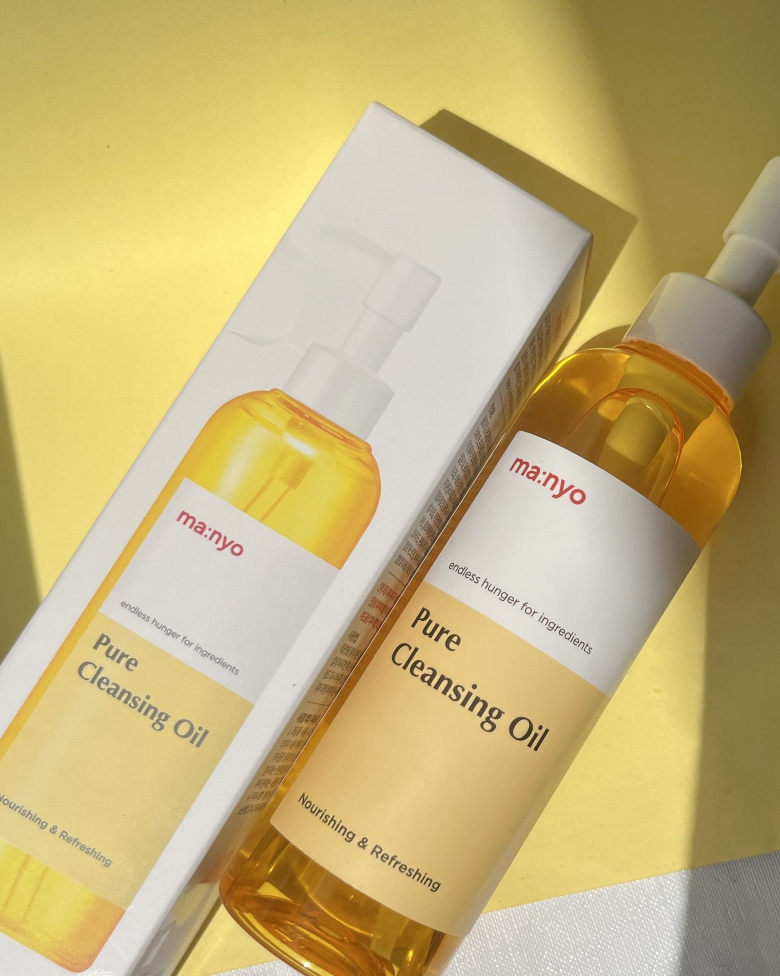ma:nyo Pure Cleansing Oil pump bottle of golden oil lying next to its white box on a sunny yellow surface.