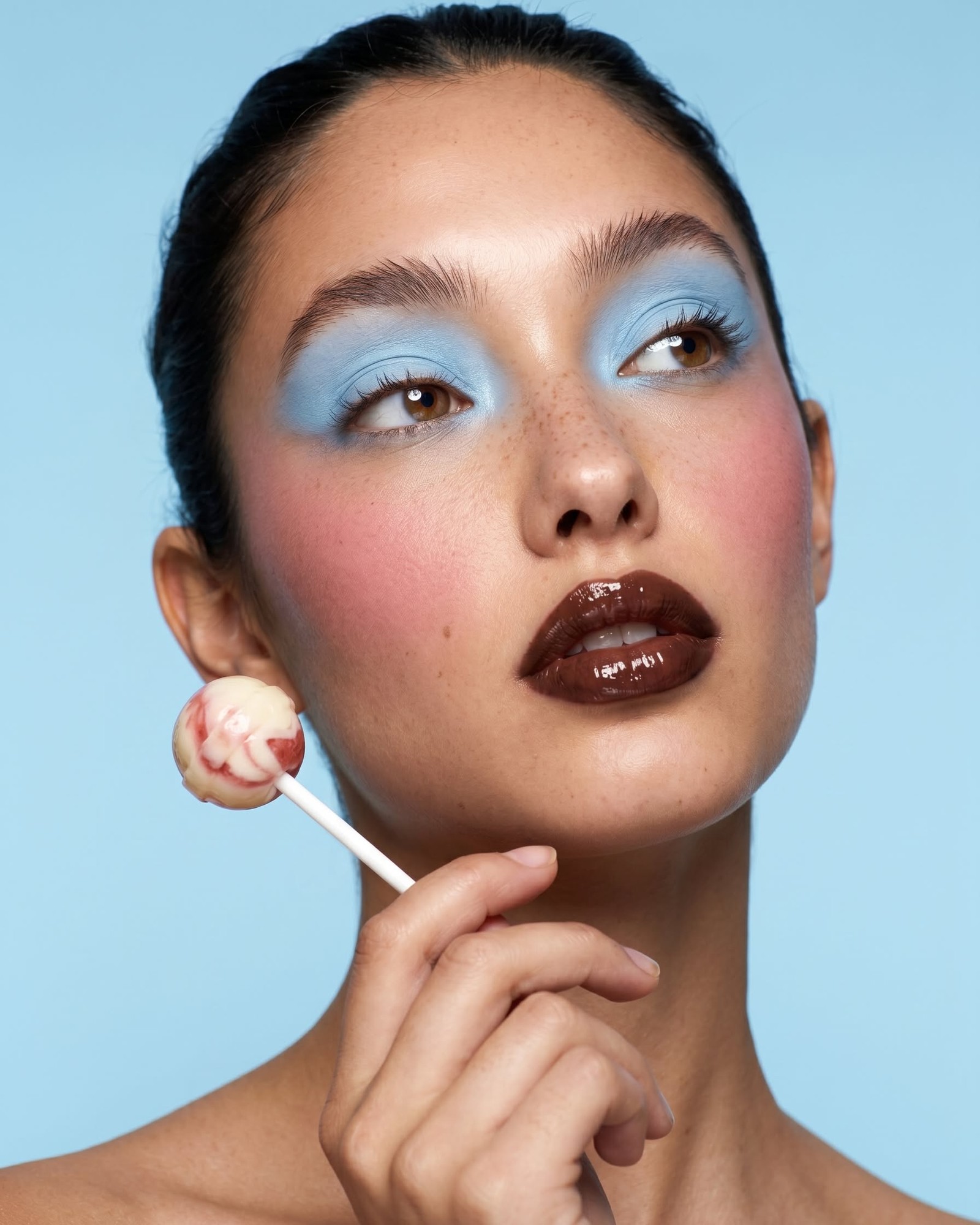 Model with slicked-back hair, pale blue eyeshadow, pink blush and a glossy chocolate-brown lip holding a swirled lollipop against a blue background