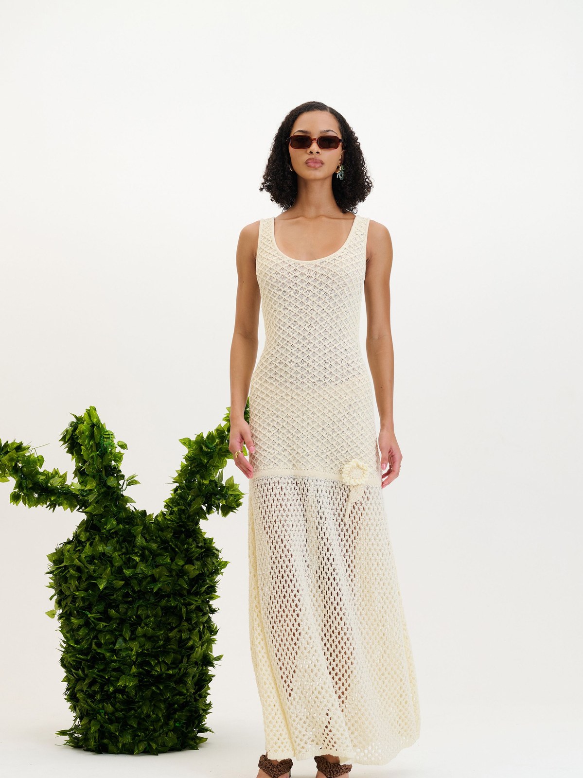 Model in an ivory open-knit dress beside a shaped green plant