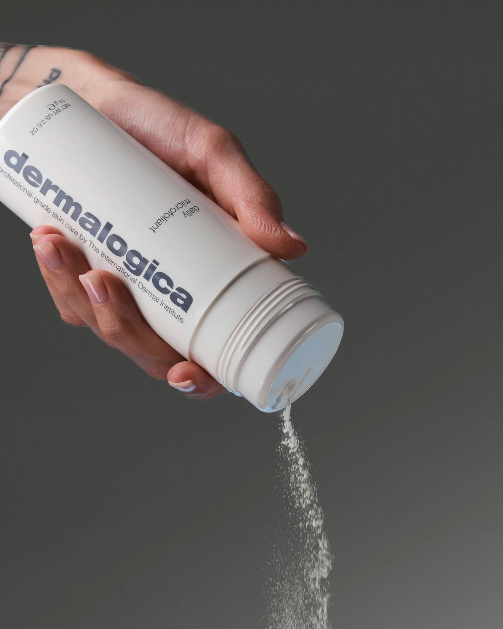 Hand tipping a white Dermalogica Daily Microfoliant jar to pour a thin stream of powder against a dark grey background