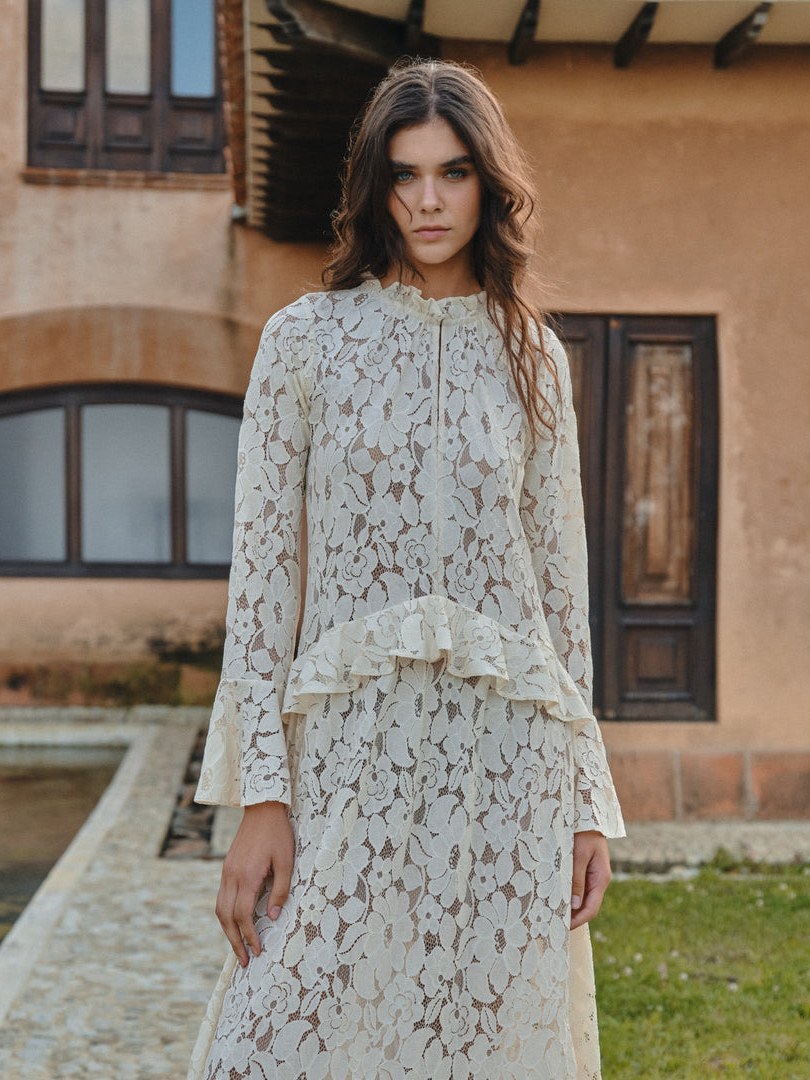 Woman in a white floral lace dress outside a stone courtyard.