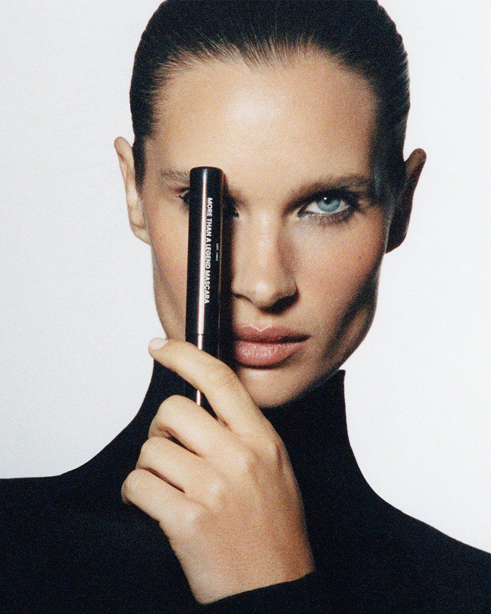 Woman with slicked-back hair in a black turtleneck holding a slim mascara tube vertically across one eye