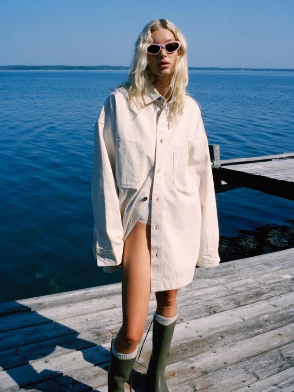 Elsa Hosk in a long white shirt on a wooden dock beside blue water