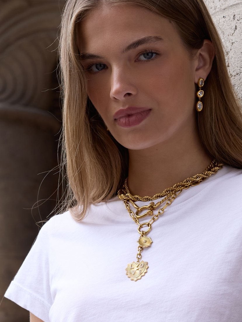 Woman in a white tee wearing layered gold chains and drop earrings against carved stone