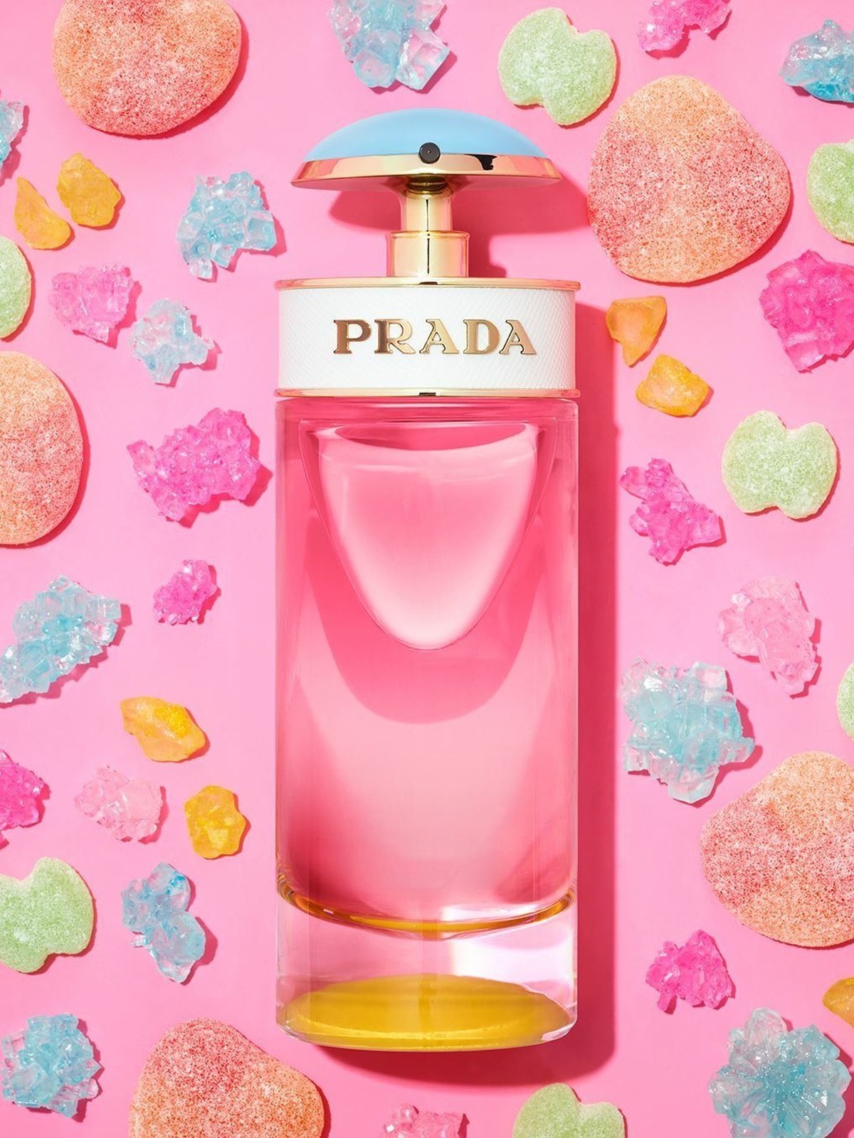Prada perfume bottle with a pink liquid, white band and pale blue cap surrounded by candy crystals and gummies on a pink background