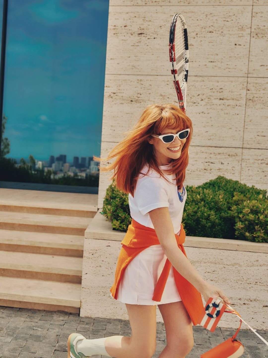 Natasha Lyonne in white with an orange layer beside a coral suitcase