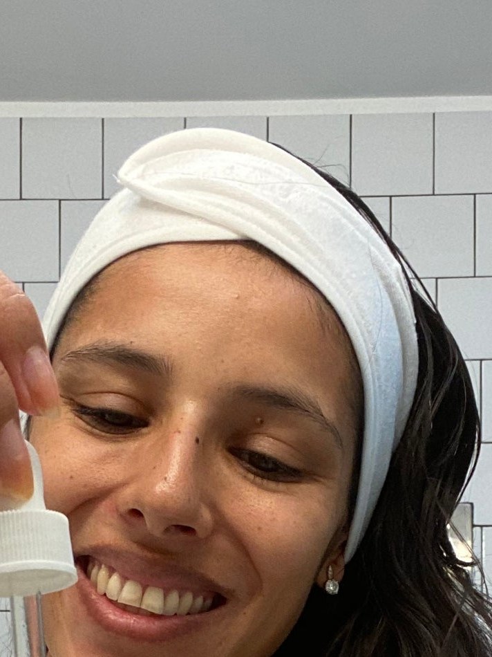 A woman applies a dropper to her hair in a tiled bathroom as part of a Ceremonia haircare ritual.