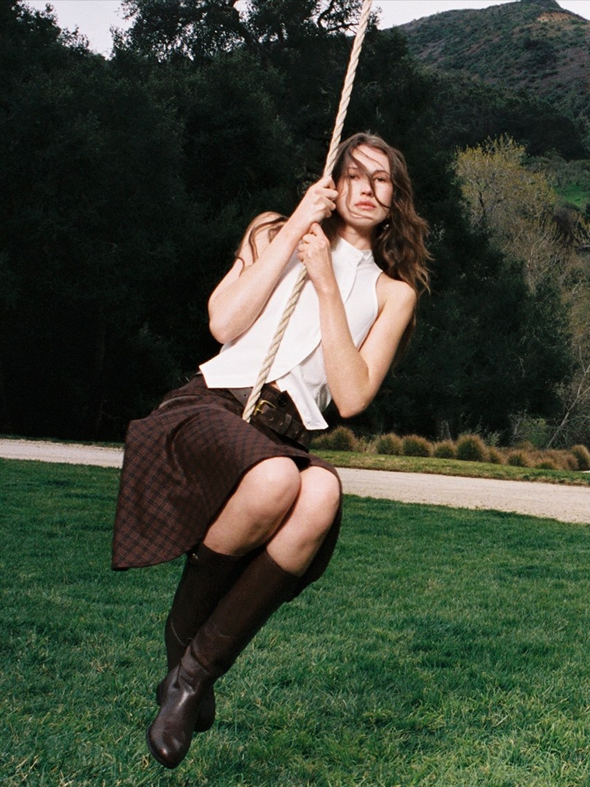 Model in a white top and dark skirt holding a rope swing in a green landscape