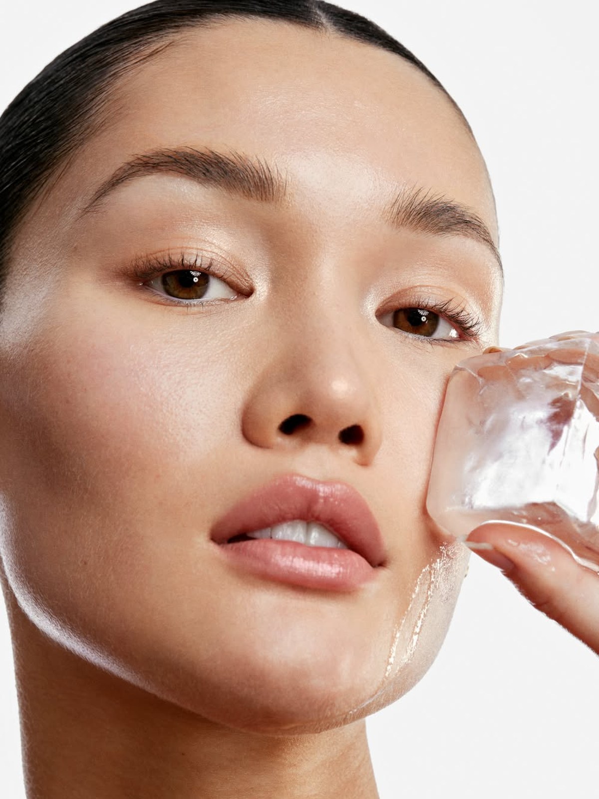 Woman with slicked-back dark hair holding a clear ice cube against her cheek on a white background