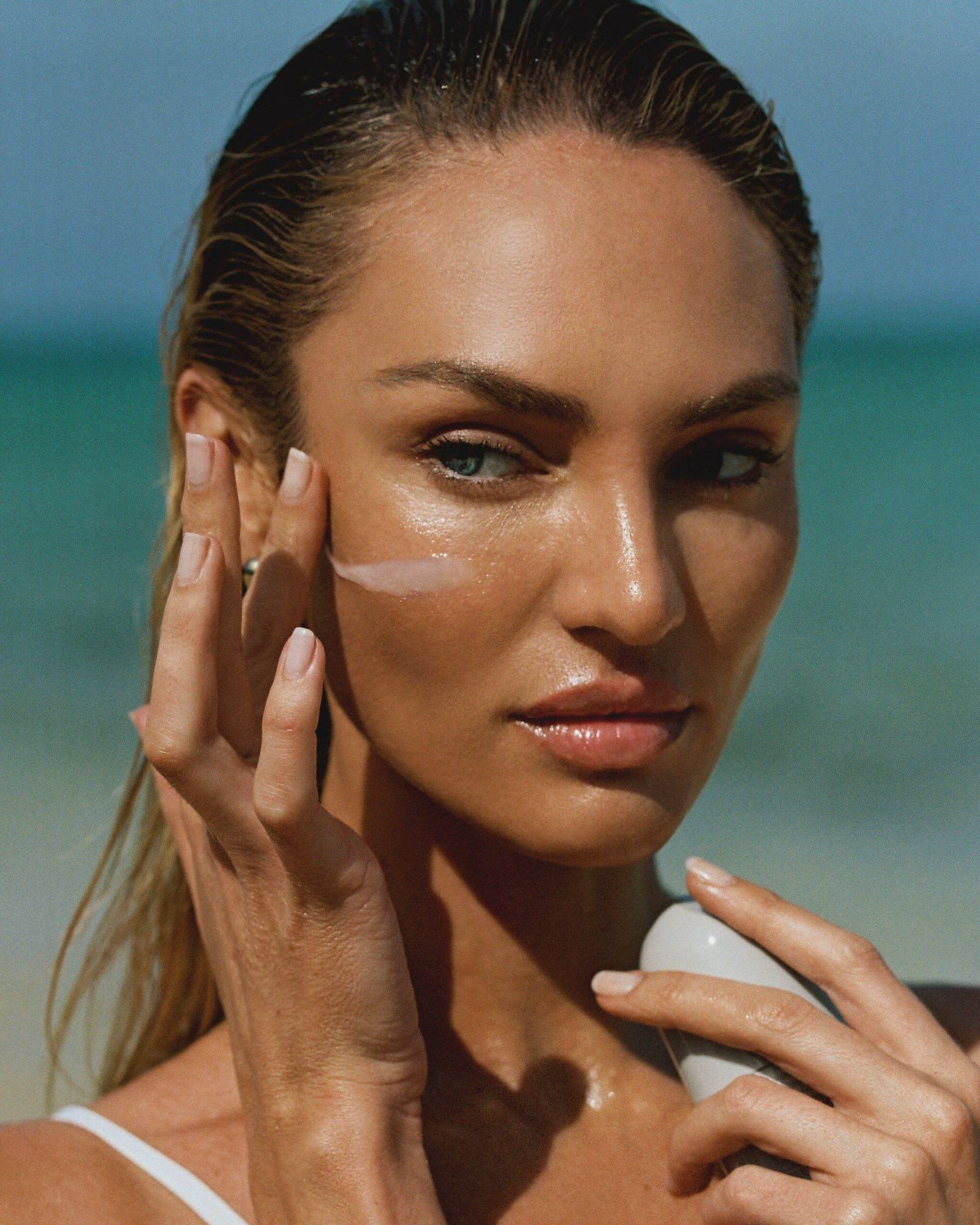 Woman with wet slicked-back hair on a beach holding a white sunscreen bottle and touching a streak of sunscreen on her cheek