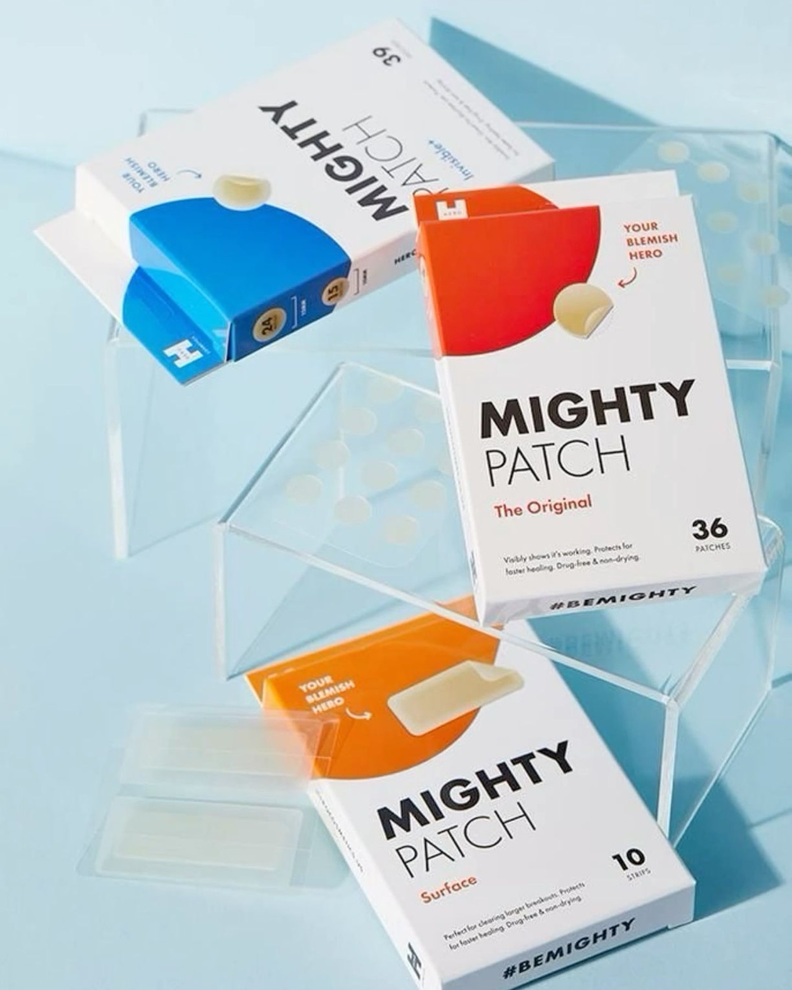 Boxes of Hero Mighty Patch Original, Invisible+ and Surface arranged on clear acrylic blocks against a pale blue background