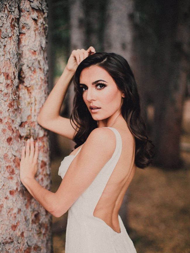 Katie May bride in a lace gown looking over her shoulder beside a tree in a forest