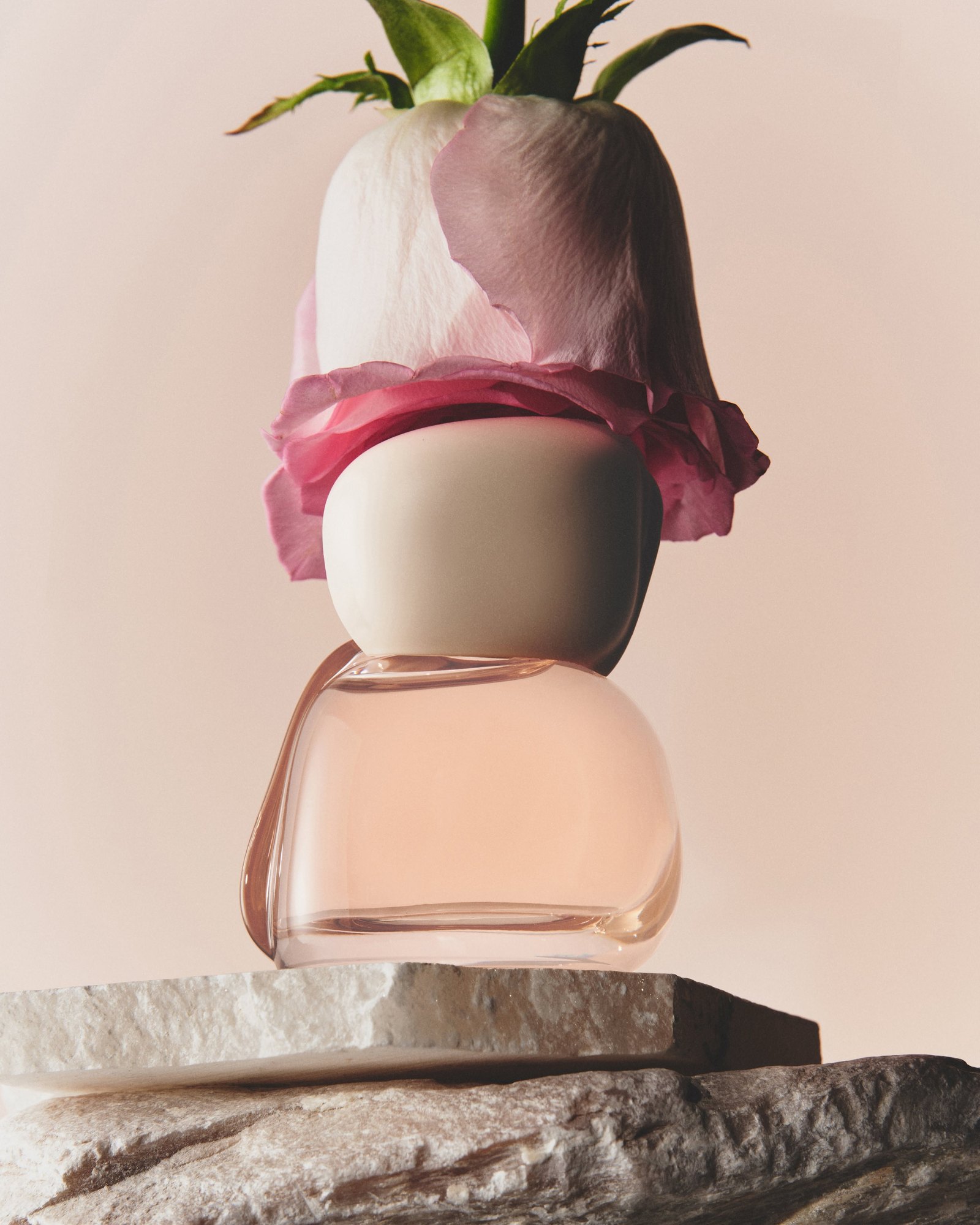 Rounded pink glass perfume bottle with a cream cap topped by a pink rose bloom on a pale stone slab