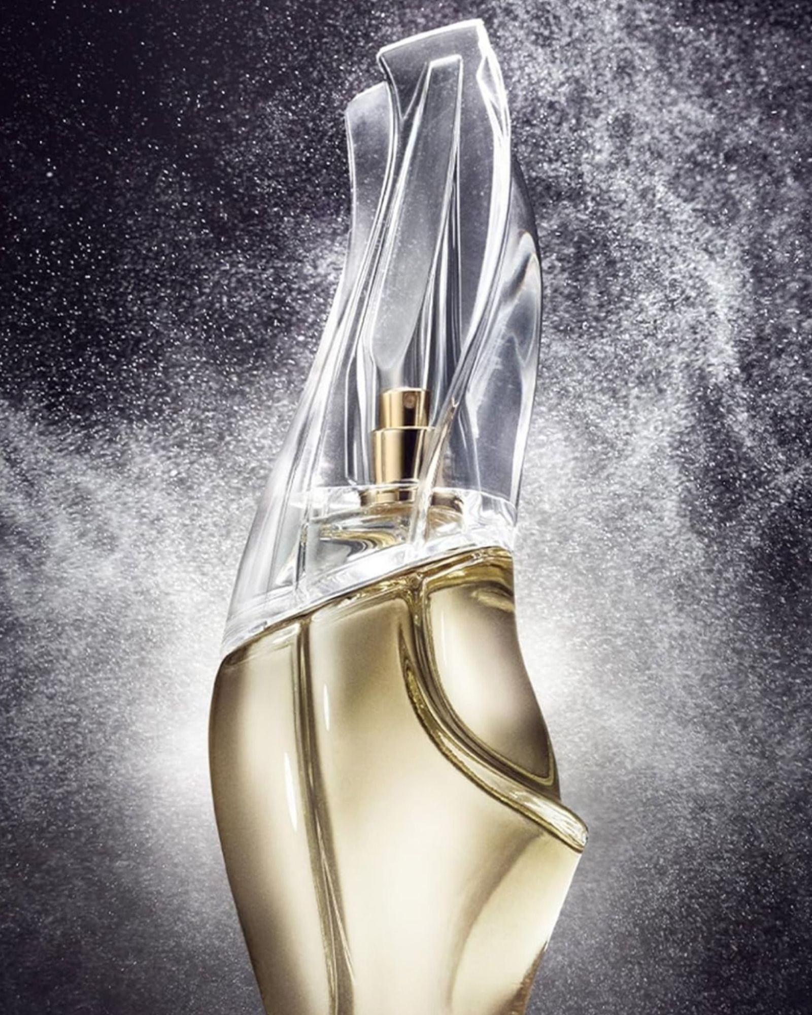 Sculptural Cashmere Mist perfume bottle with a glass top and pale gold liquid, surrounded by a cloud of fine spray against a dark background