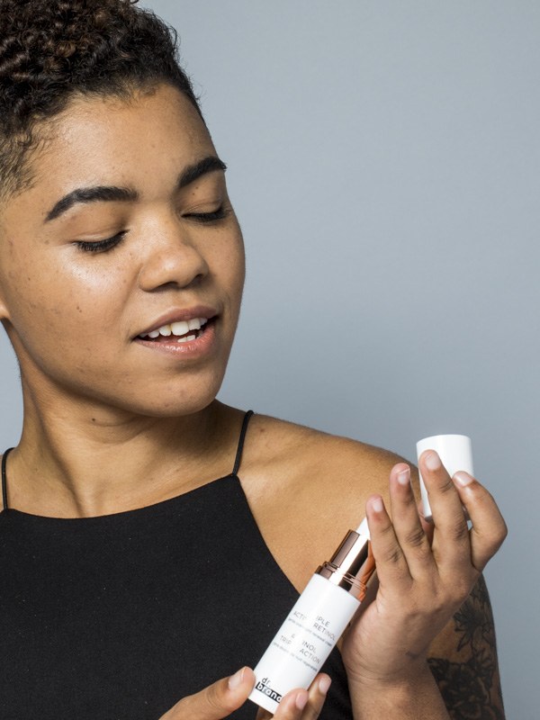 Model in a black top studies a white skincare bottle against a cool gray studio background.