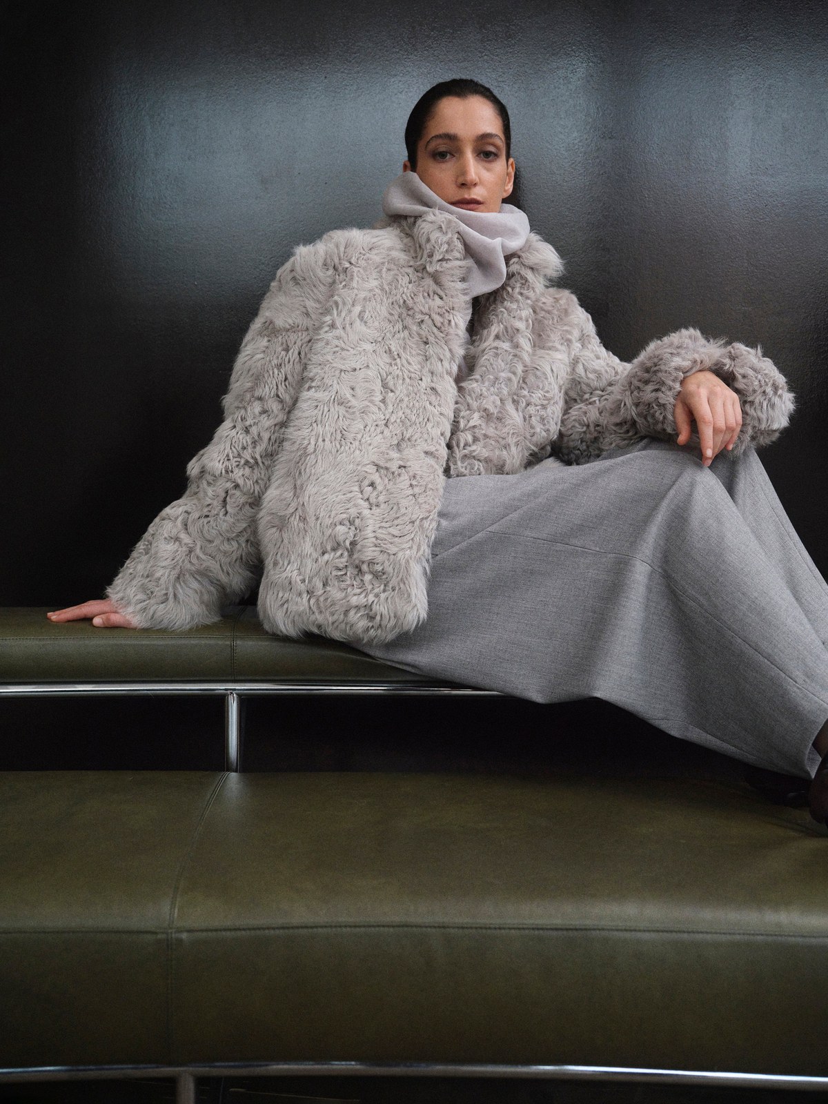 Model in a textured grey coat and draped trousers on a dark studio sofa