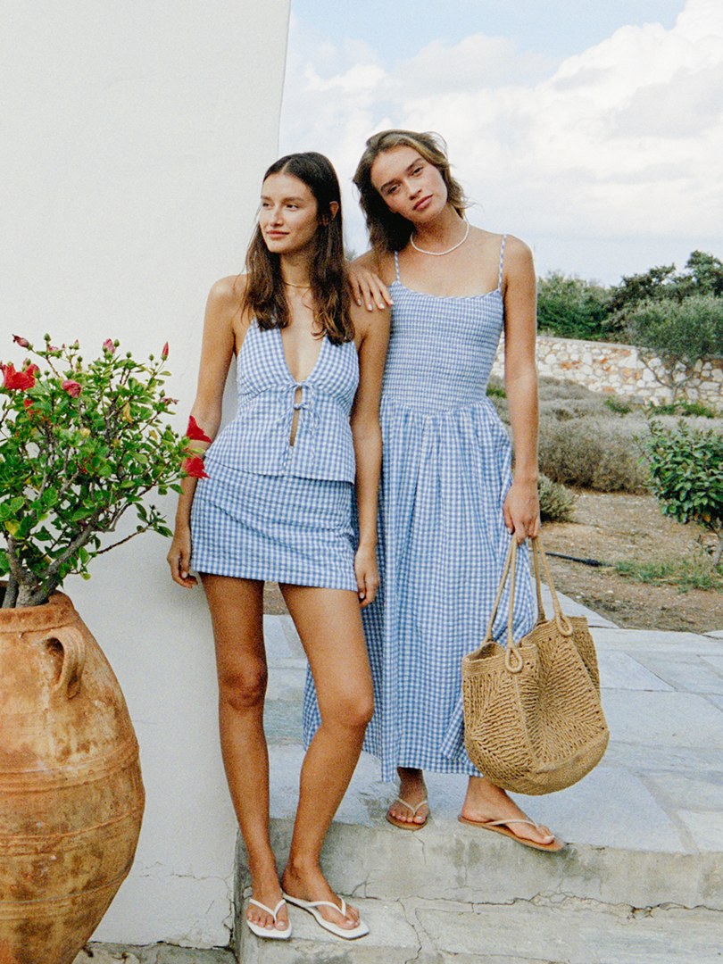Two models in blue gingham dresses carrying woven totes in a coastal courtyard