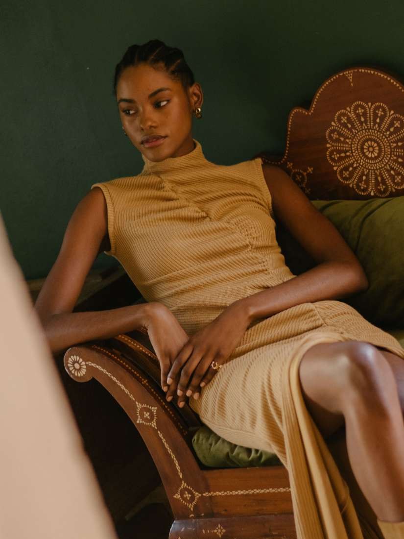 Model seated in a tan textured dress on an embroidered wooden chair