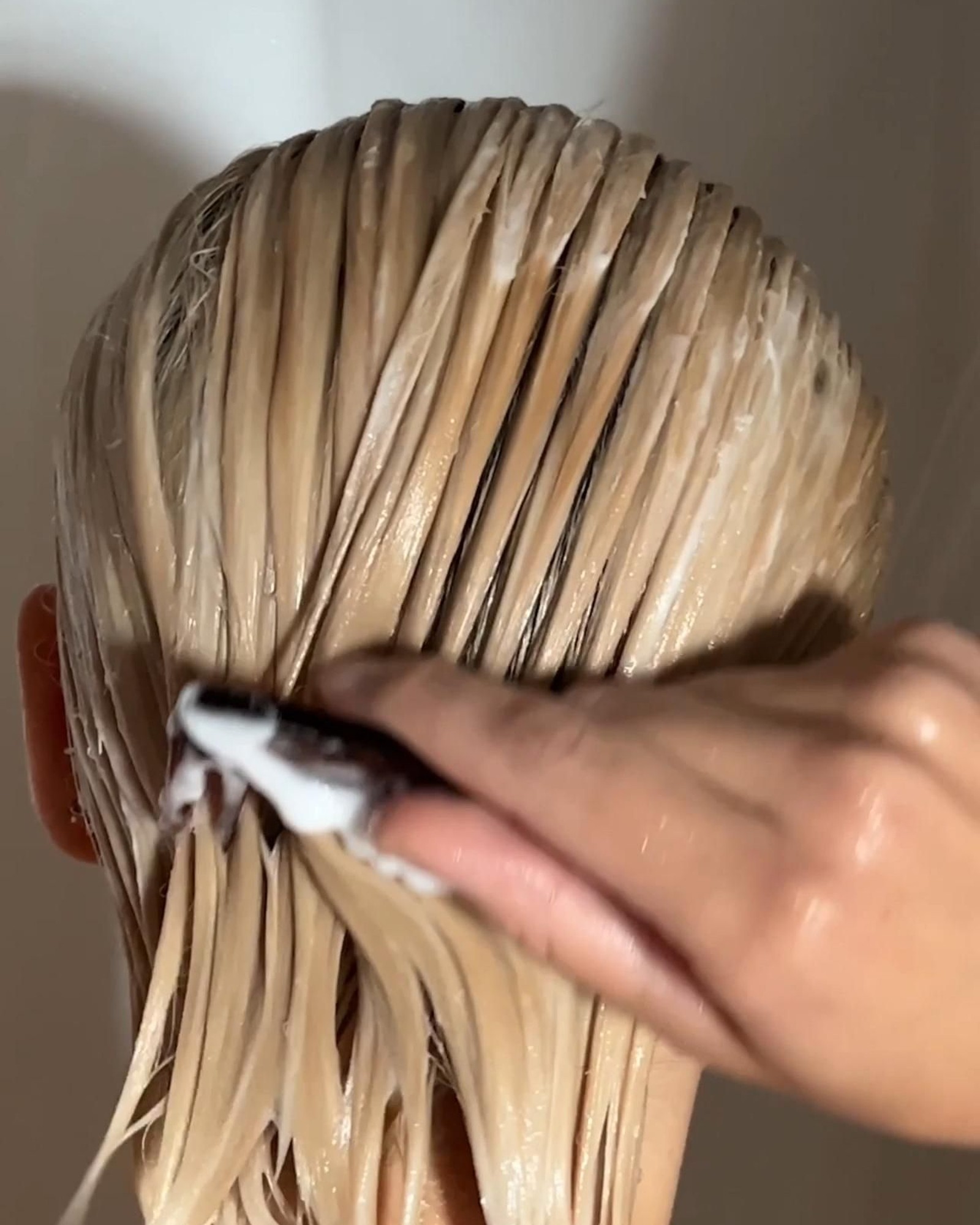 Back of a head with wet blonde hair slicked down as a hand smooths white conditioner through the lengths