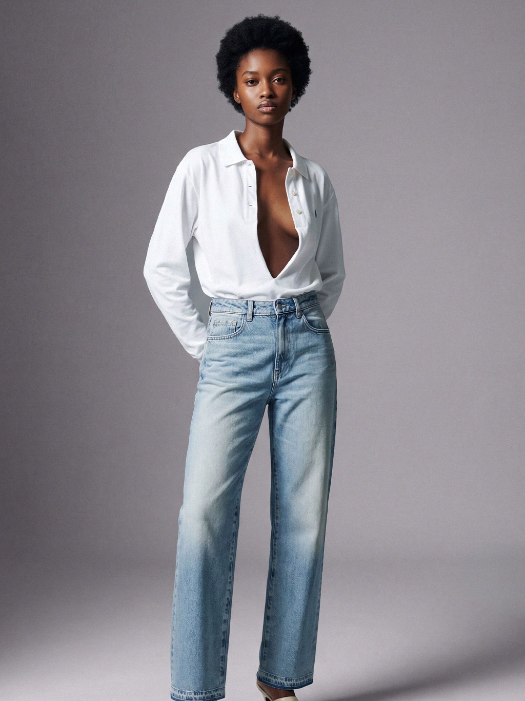 Model in straight light-wash jeans and a white blouse against a gray studio backdrop.