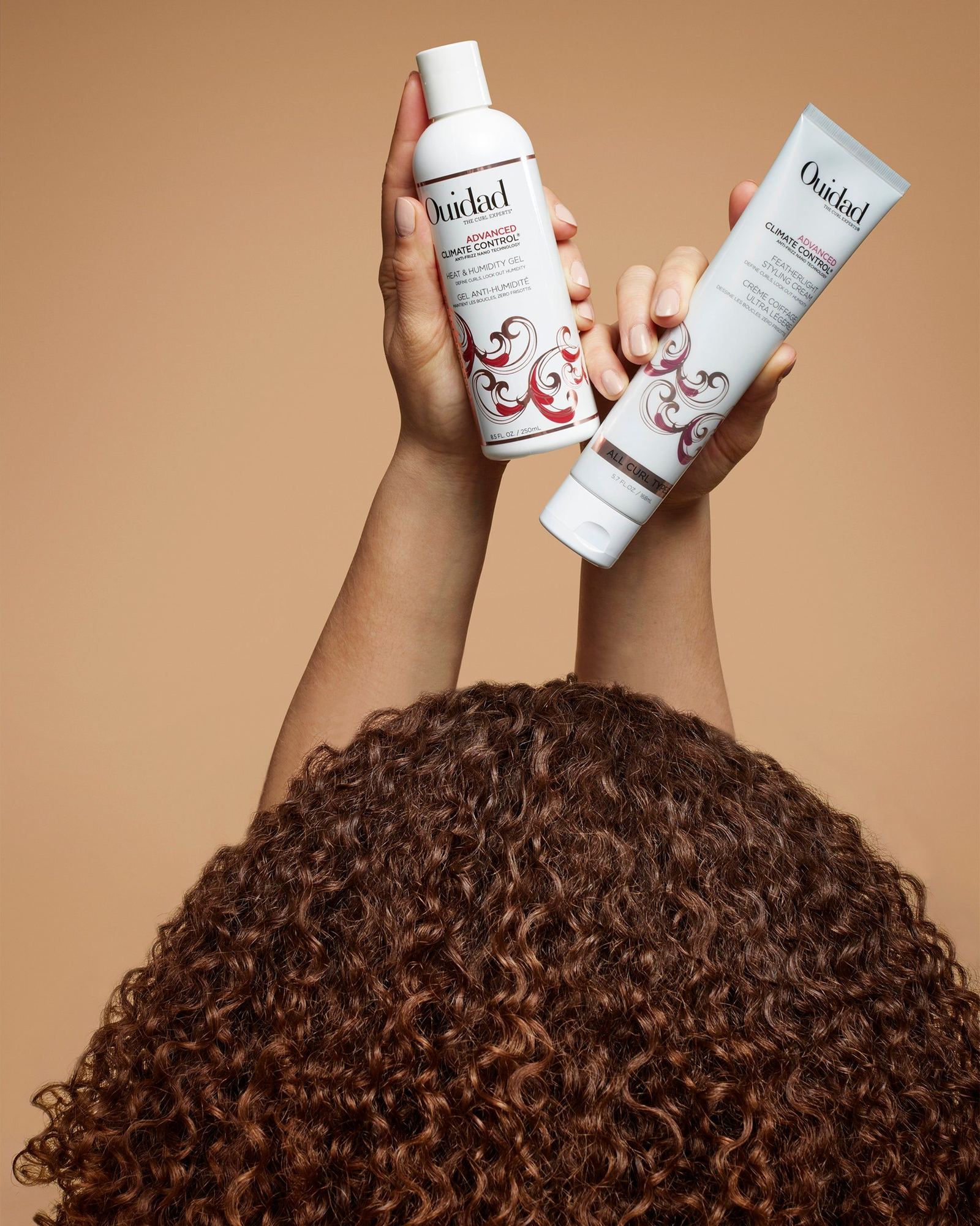 Two hands raised holding a Climate Control Heat and Humidity Gel bottle and a Featherlight Styling Cream tube above a head of brown curls on a tan background