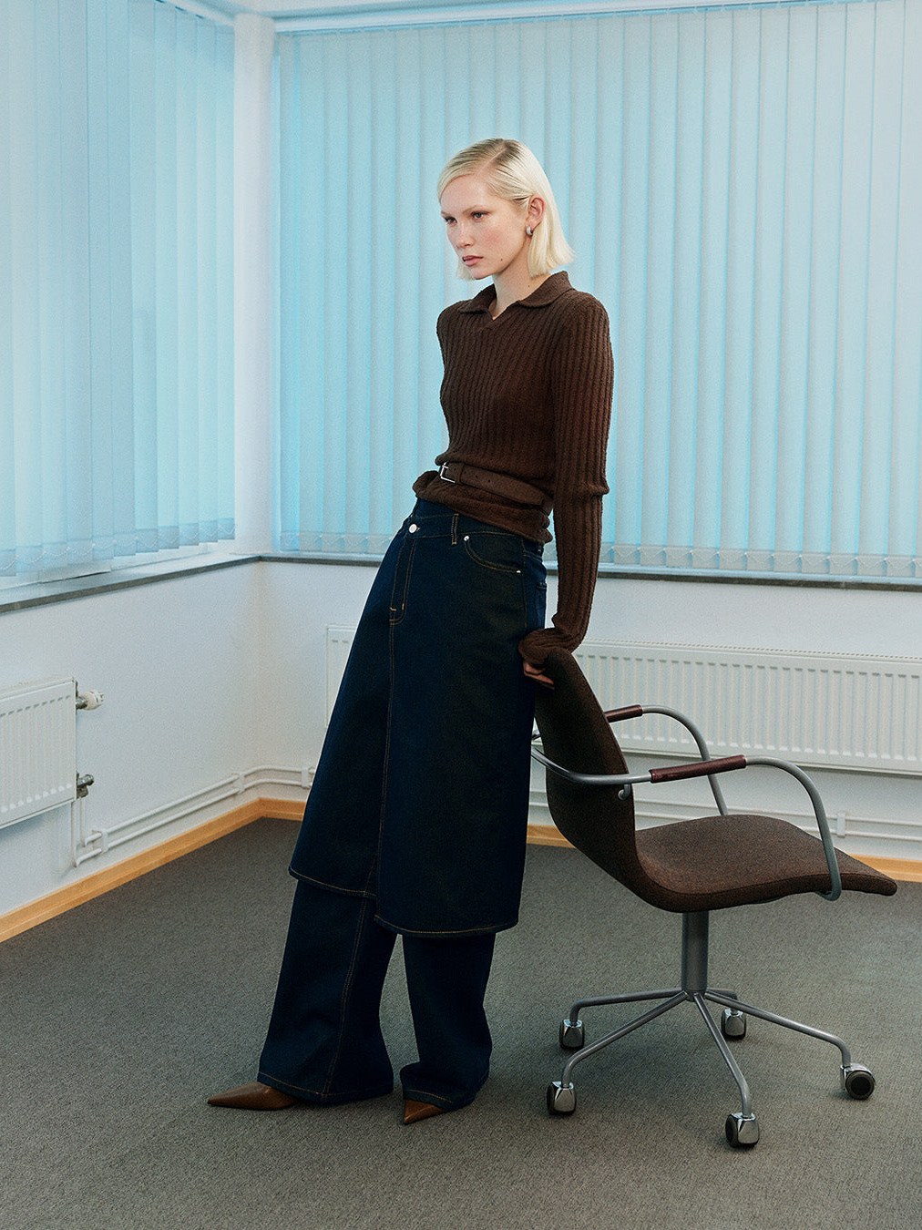 Model in a dark knit top and long denim skirt against pale blue blinds