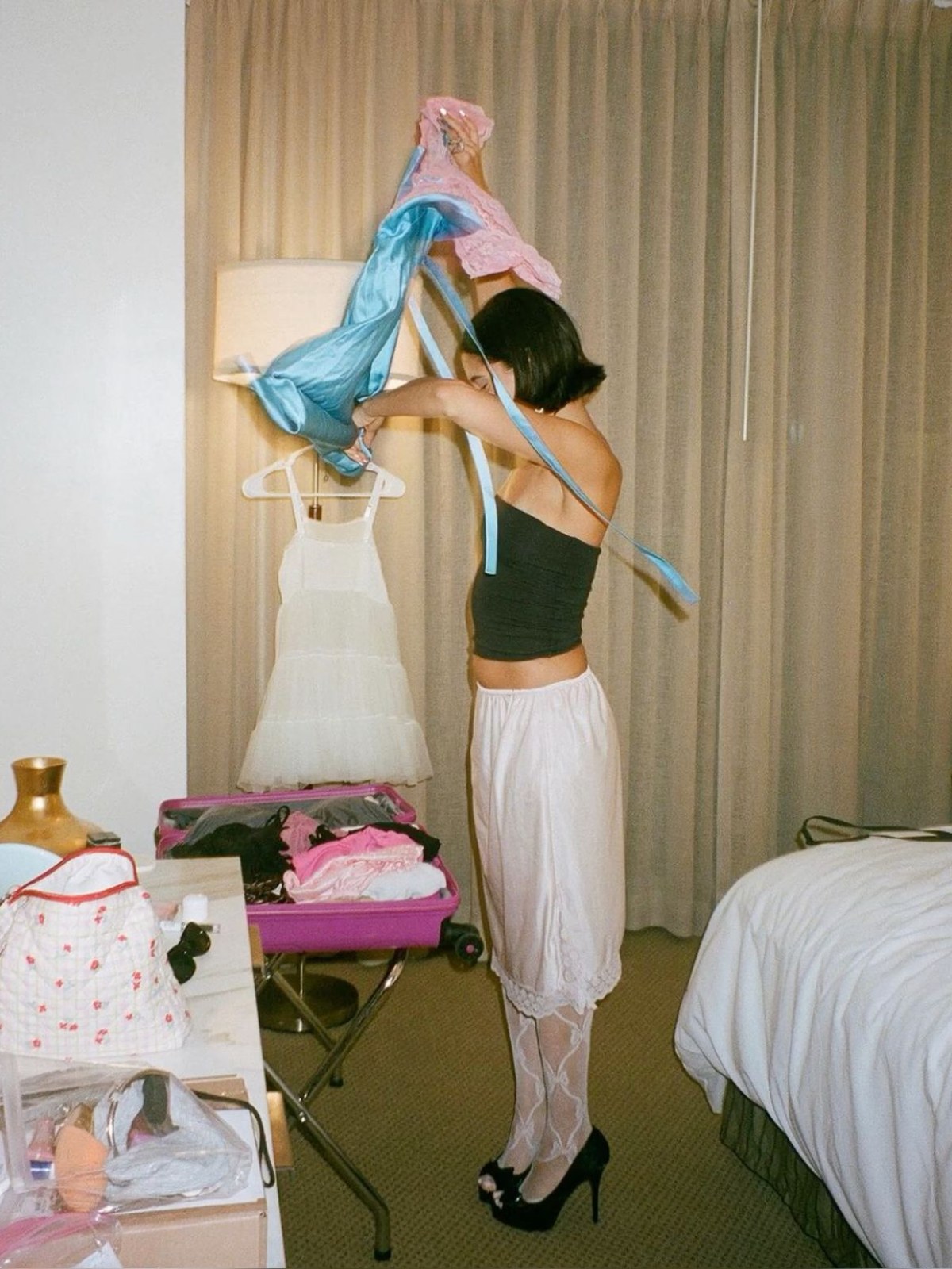 A woman pulls on a top in a bedroom strewn with clothes