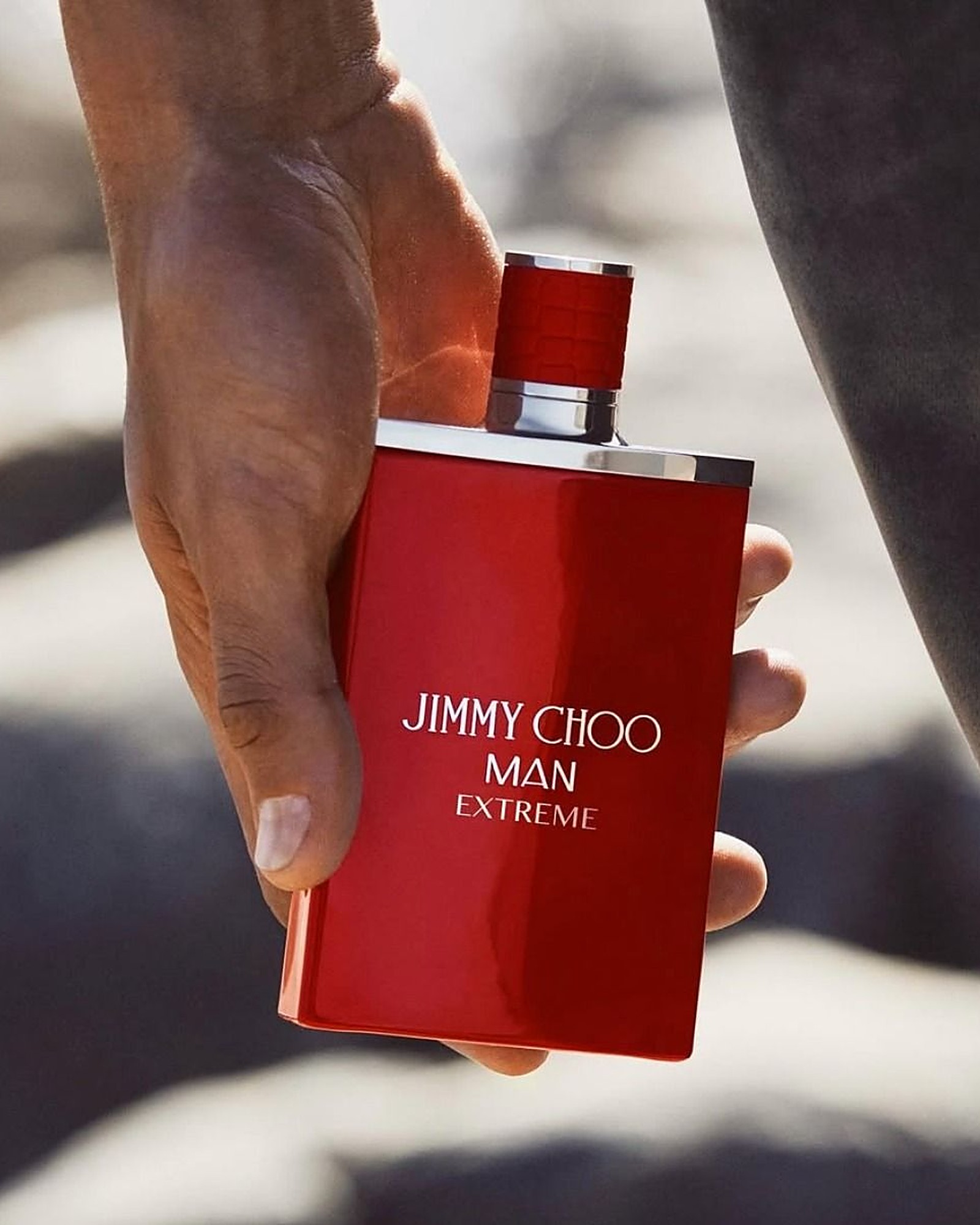 Hand holding a red Jimmy Choo Man Extreme bottle against a soft blurred rocky background