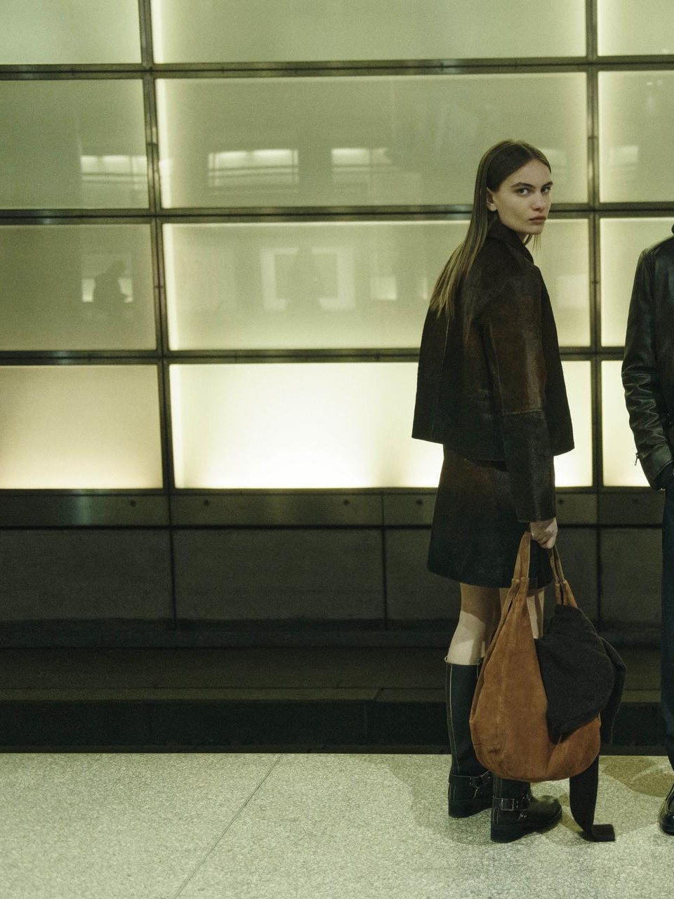 Woman in a dark brown coat and boots carrying a large bag beneath illuminated Berlin windows