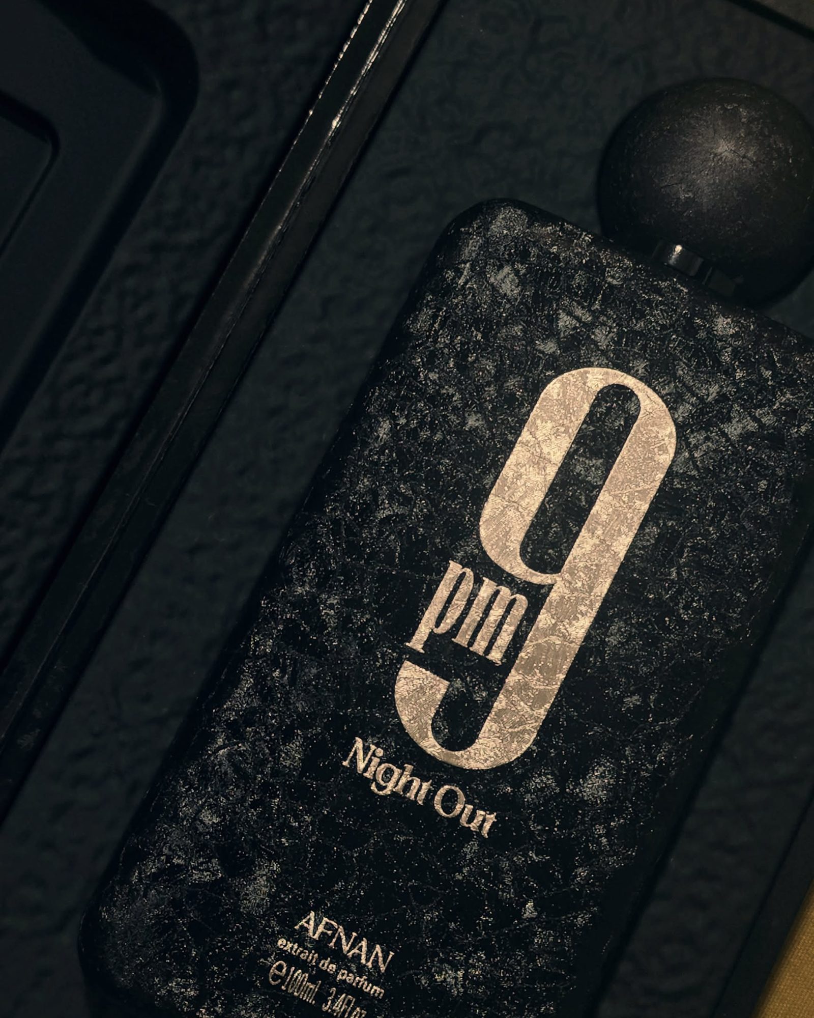 Close-up of the Afnan 9pm Night Out bottle, textured charcoal glass with a gold 9 pm and a dark sphere cap, resting in its black box