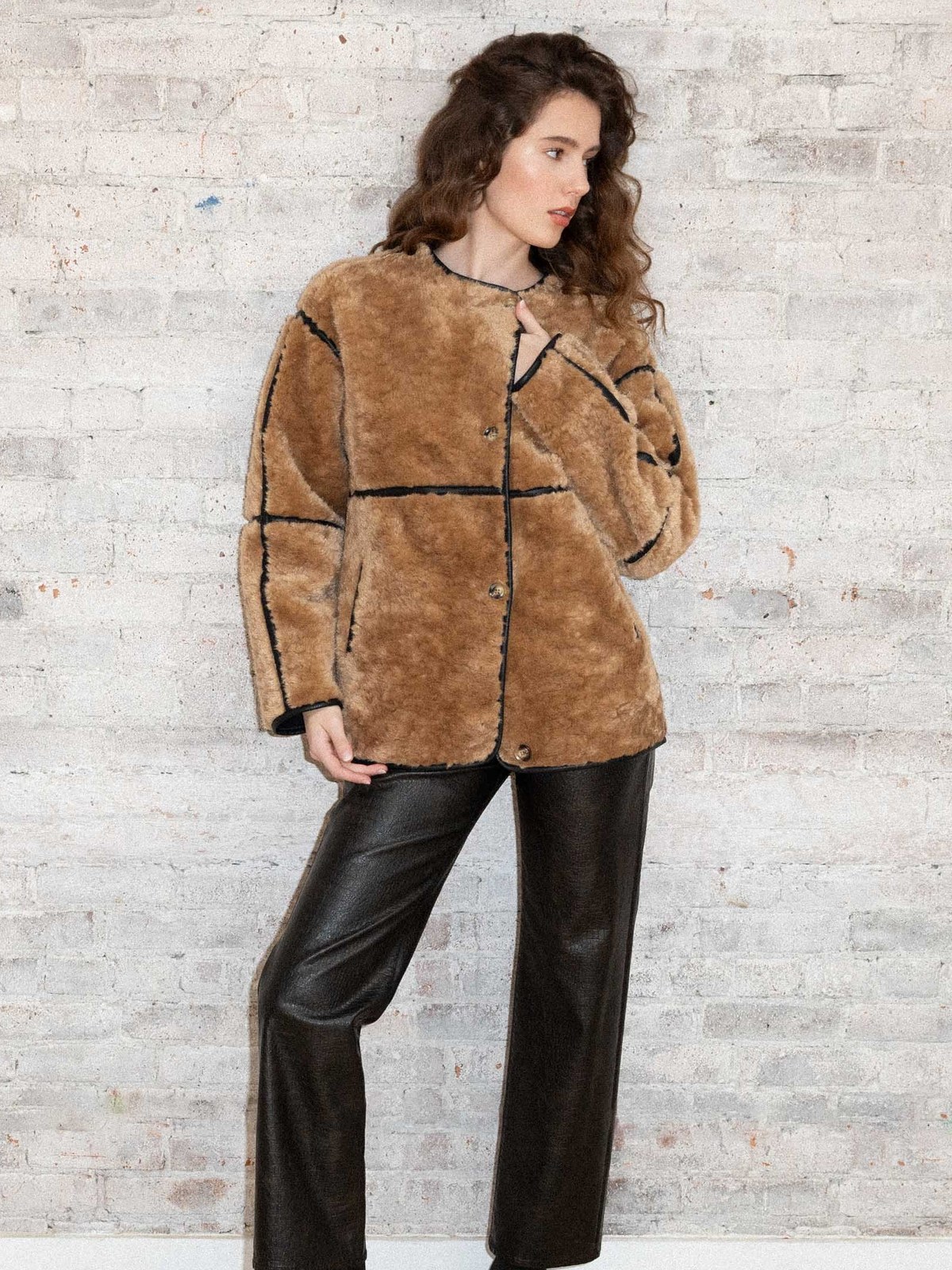 Model in a tan faux fur jacket and black trousers against pale brick