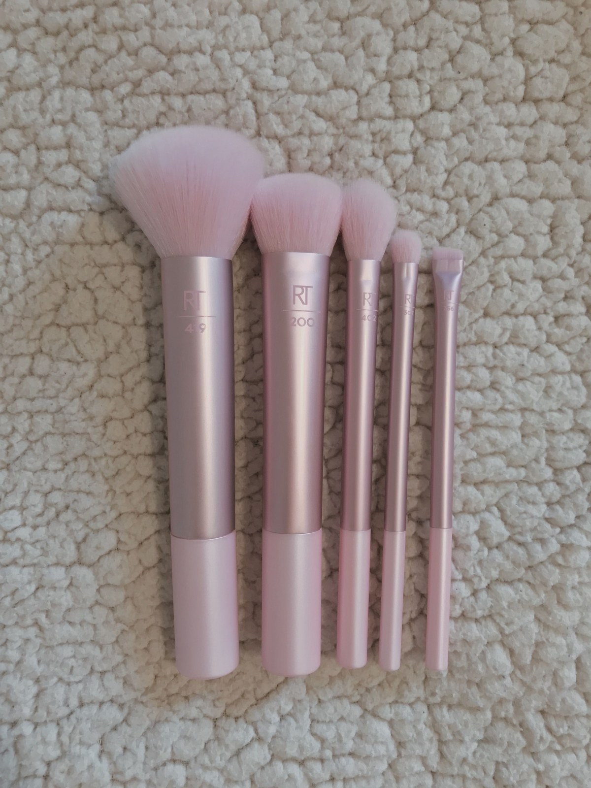 Five pink makeup brushes of varying sizes laid out on a cream rug