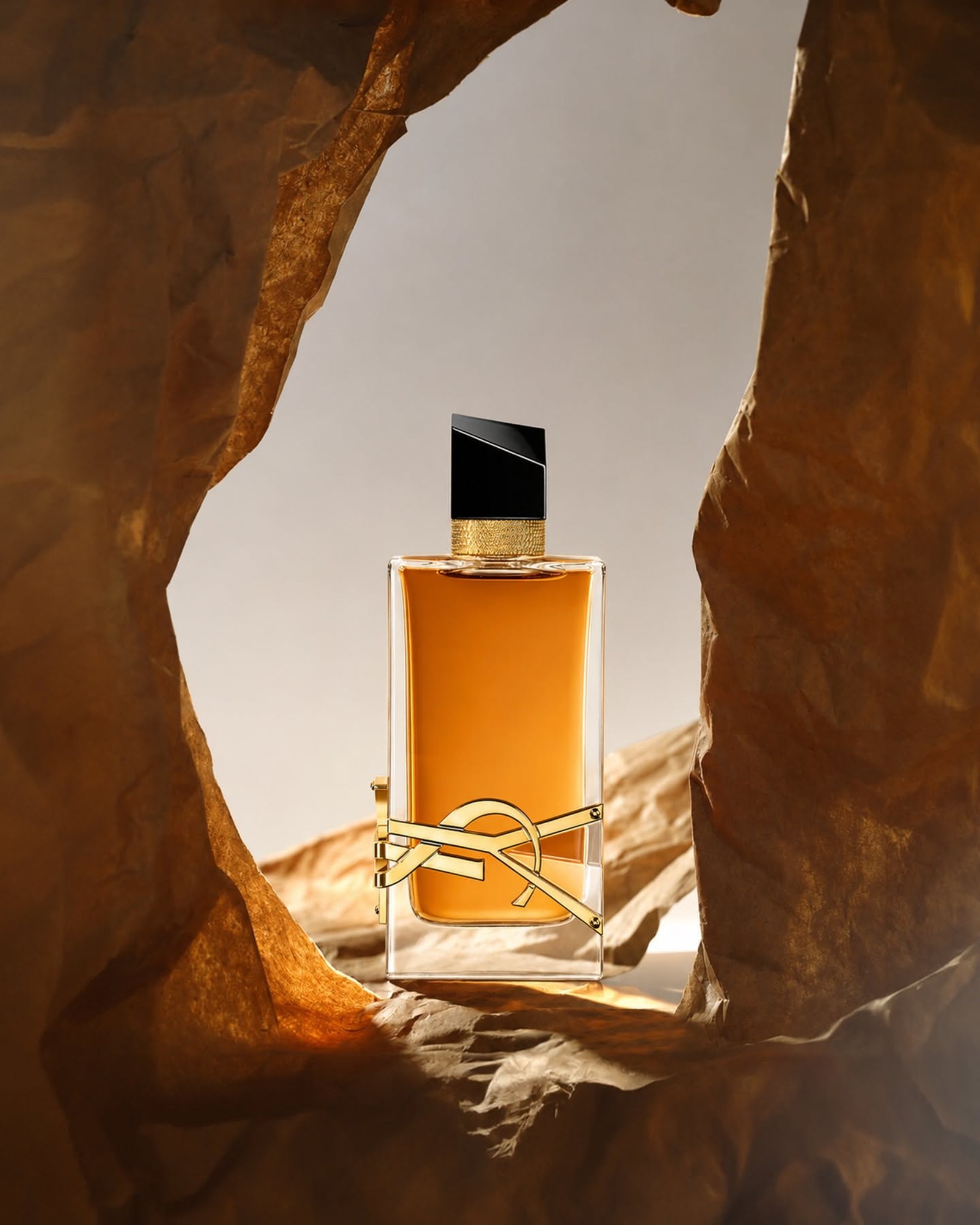 A bottle of YSL Libre Intense with amber liquid and a gold YSL emblem standing in a gap between crumpled golden paper rocks