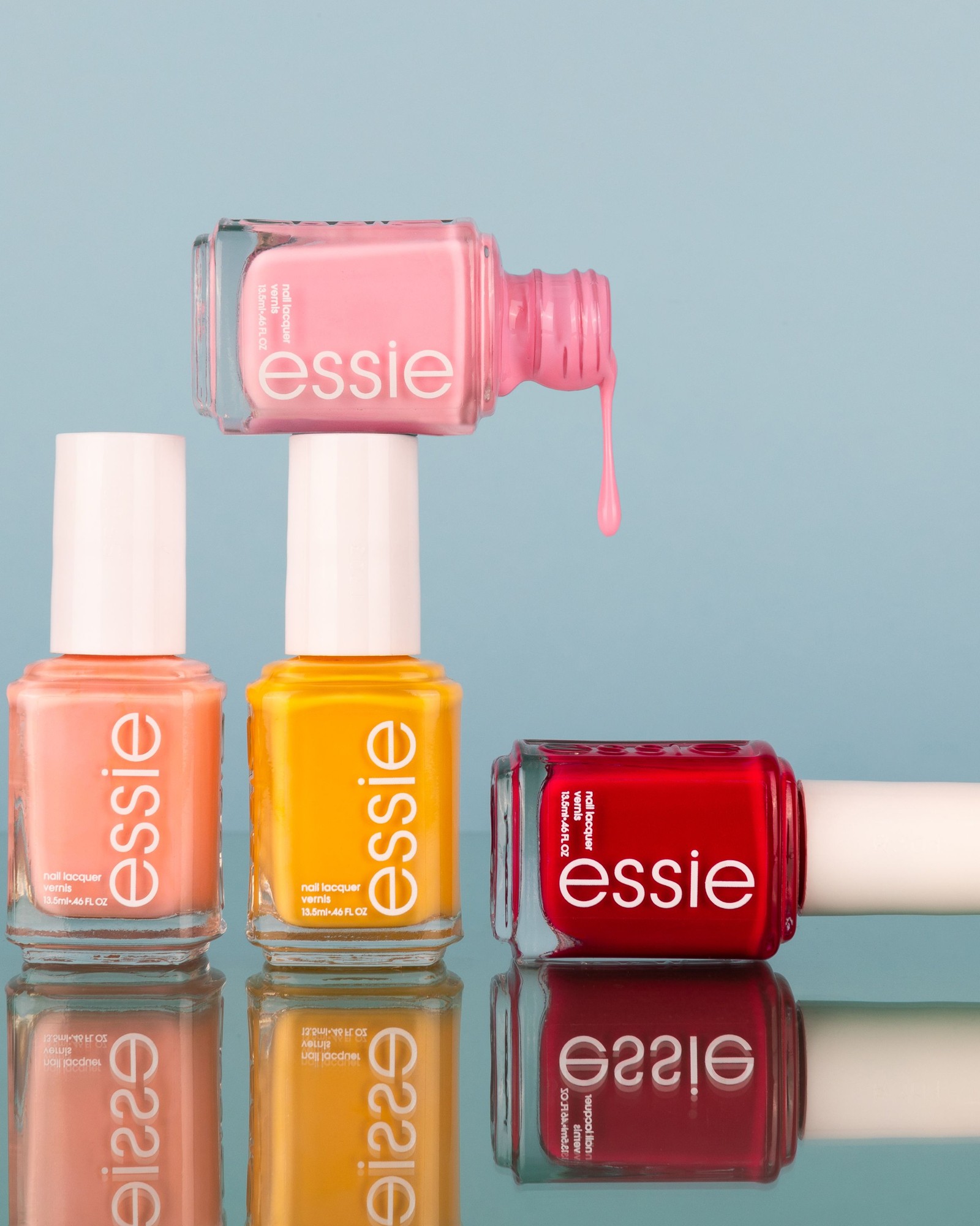 Four essie bottles in peach, marigold yellow, pink and red standing on a glossy surface against a soft blue background